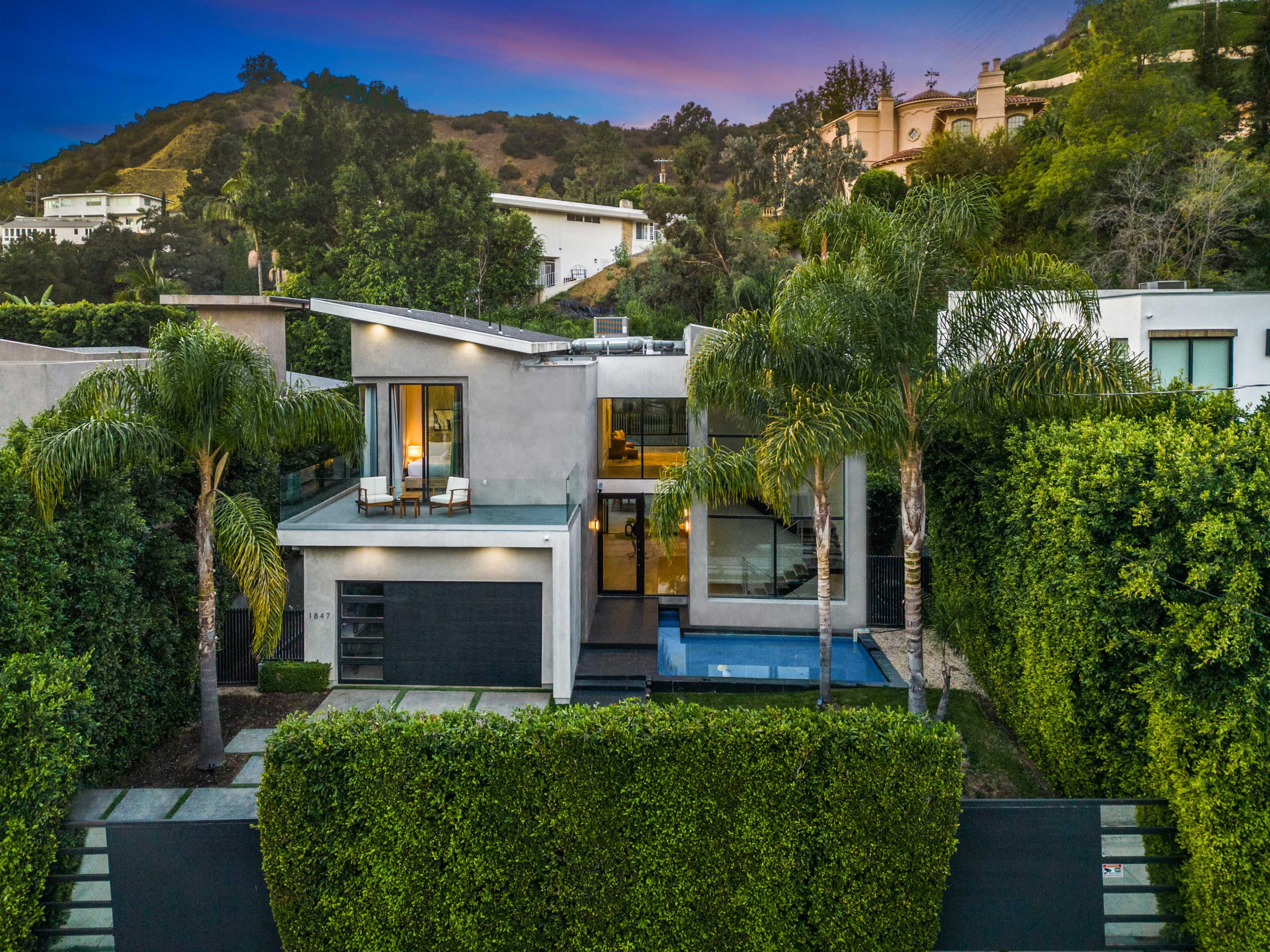 The image shows a modern two-story house surrounded by greenery and palm trees, with a driveway leading to an enclosed entrance.