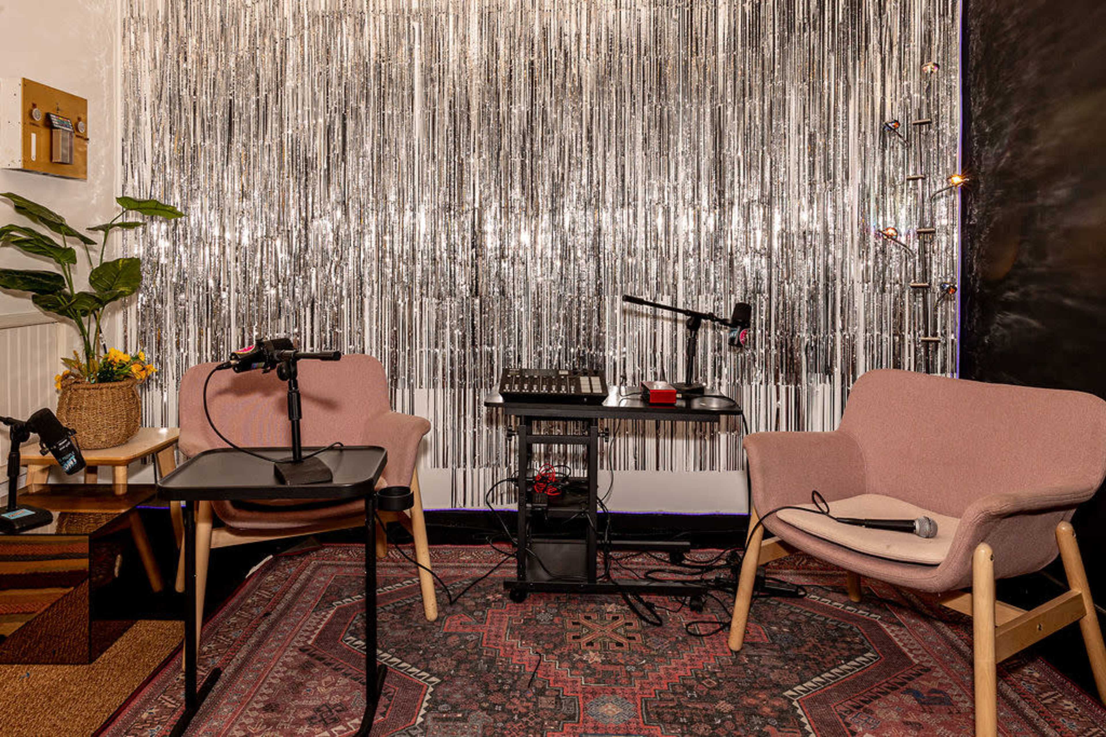 The image shows a small podcast studio with two chairs, a table, microphones, and a shiny metallic backdrop.