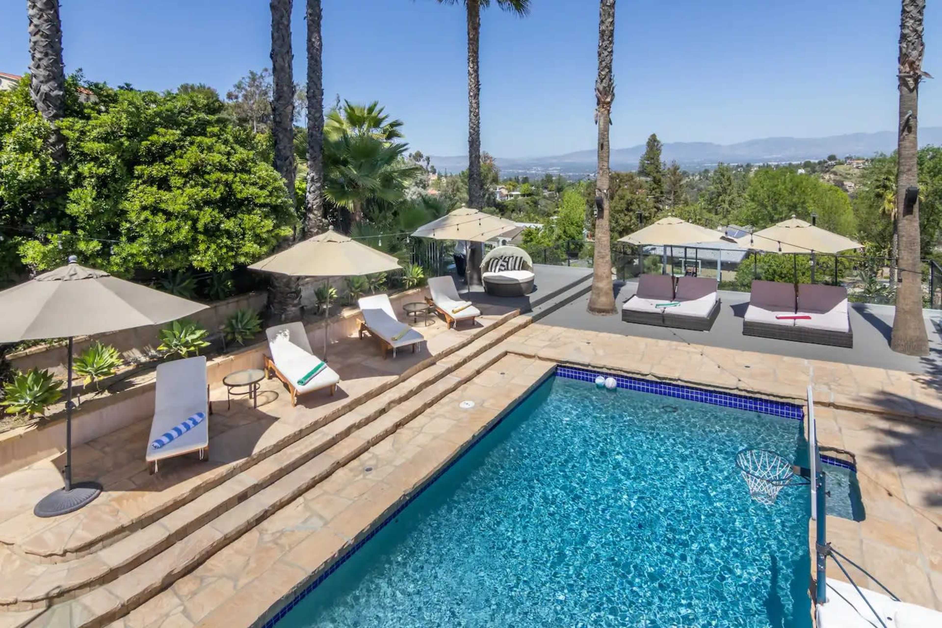 The image shows a backyard with a swimming pool surrounded by lounging chairs, umbrellas, and a view of distant hills.