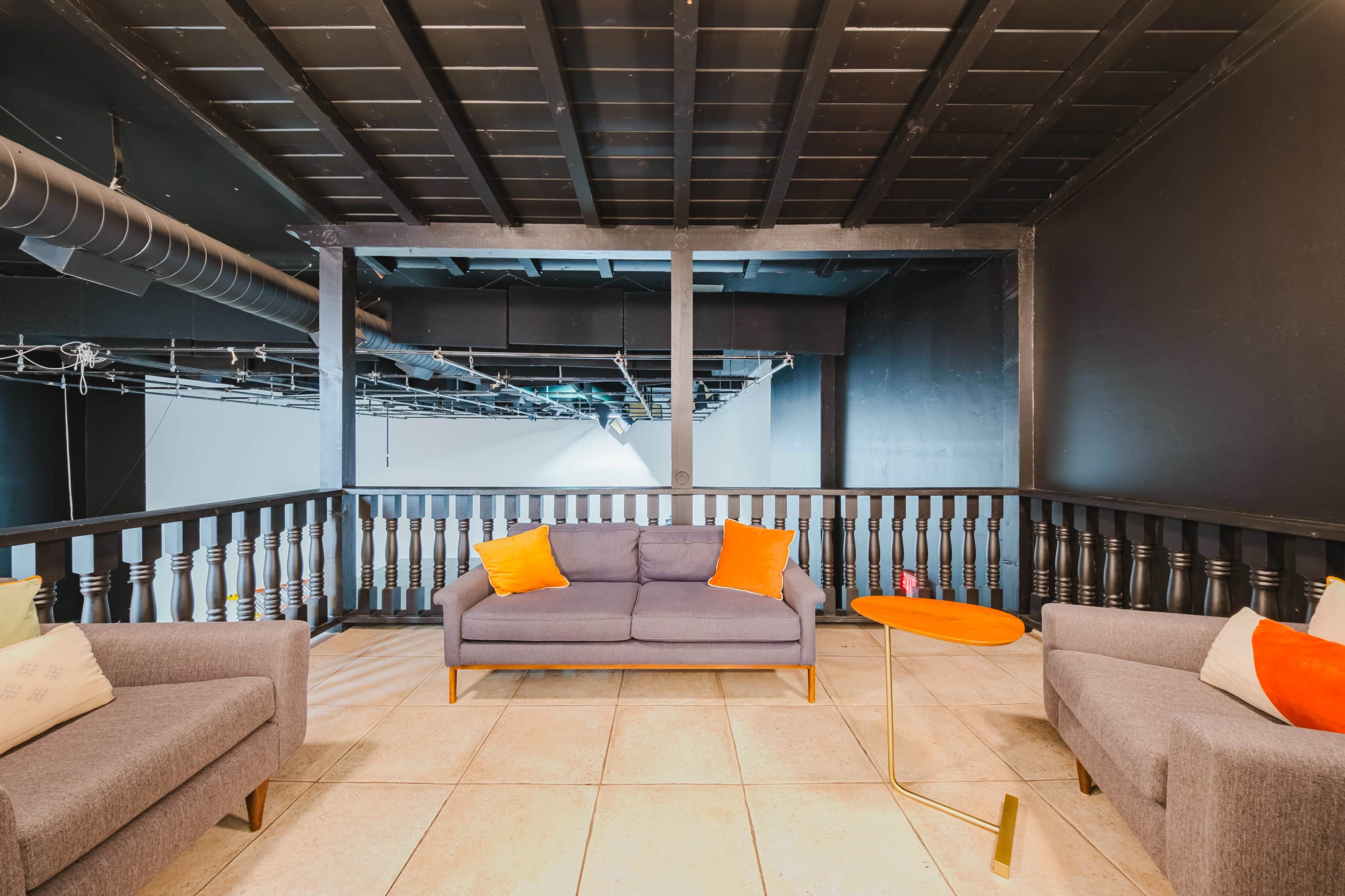 The image depicts a modern, open living area with two gray sofas, orange throw pillows, and a small orange table, set on tiled flooring beneath a black ceiling.