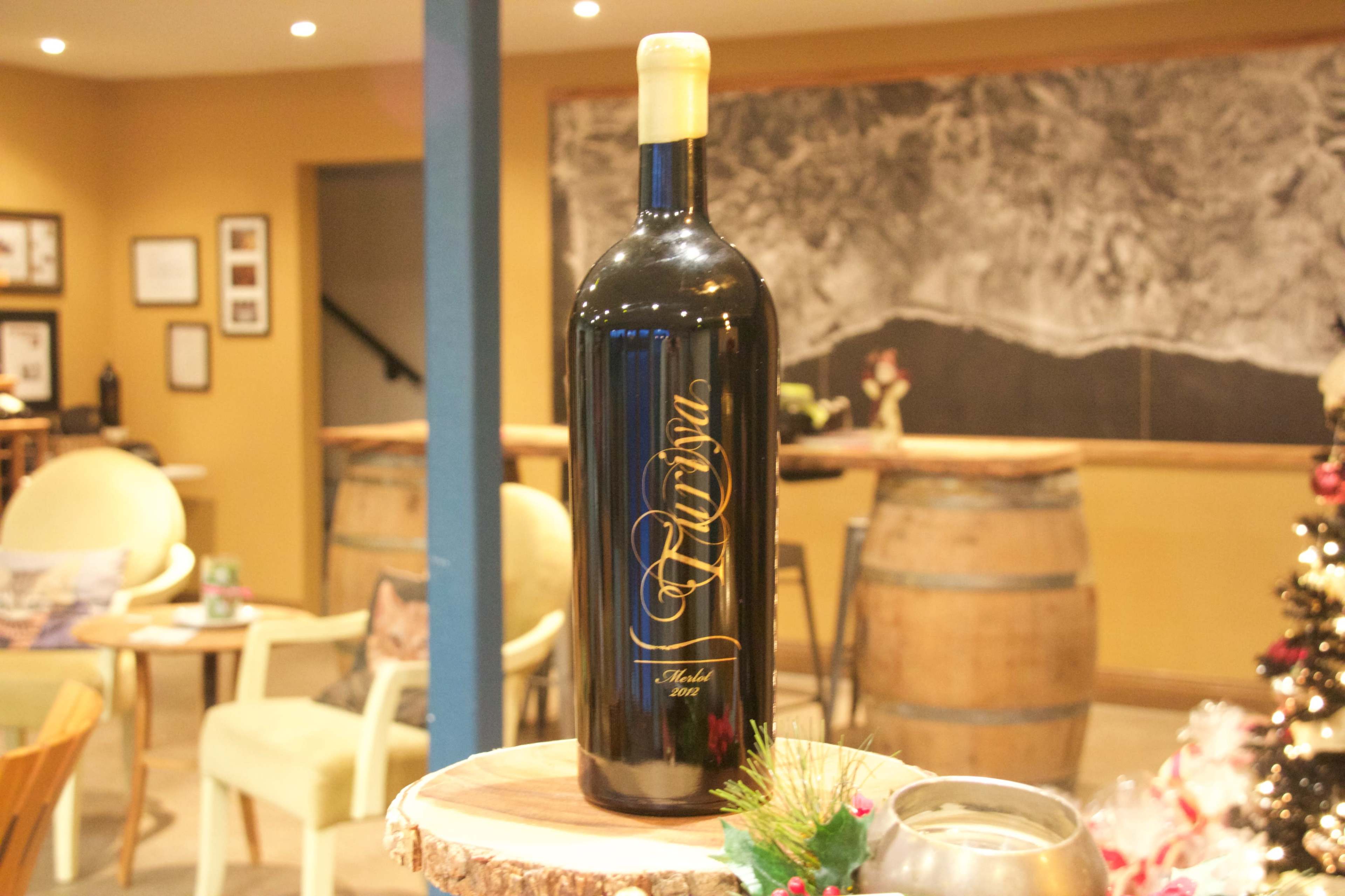 A large black wine bottle labeled "Alivio" stands on a wooden display in a warm-toned room decorated for the holidays.