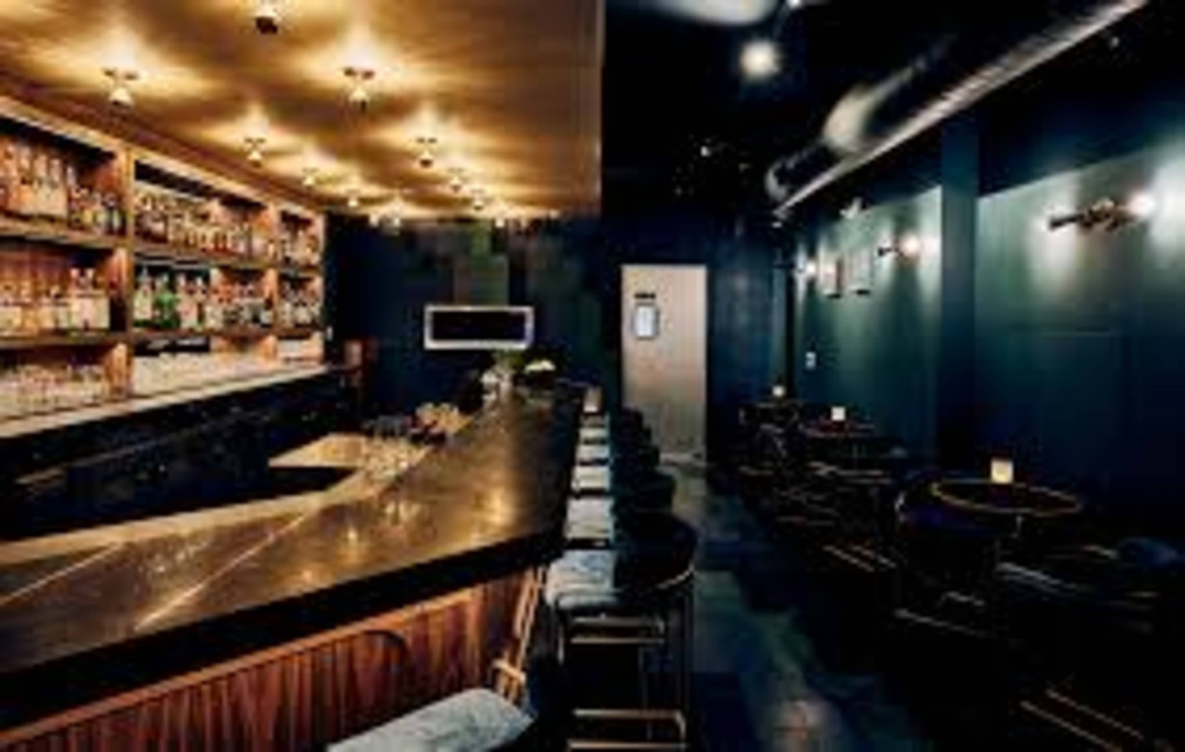 A modern bar features a sleek counter, stylish seating, and shelves stocked with various bottles of liquor under warm lighting.