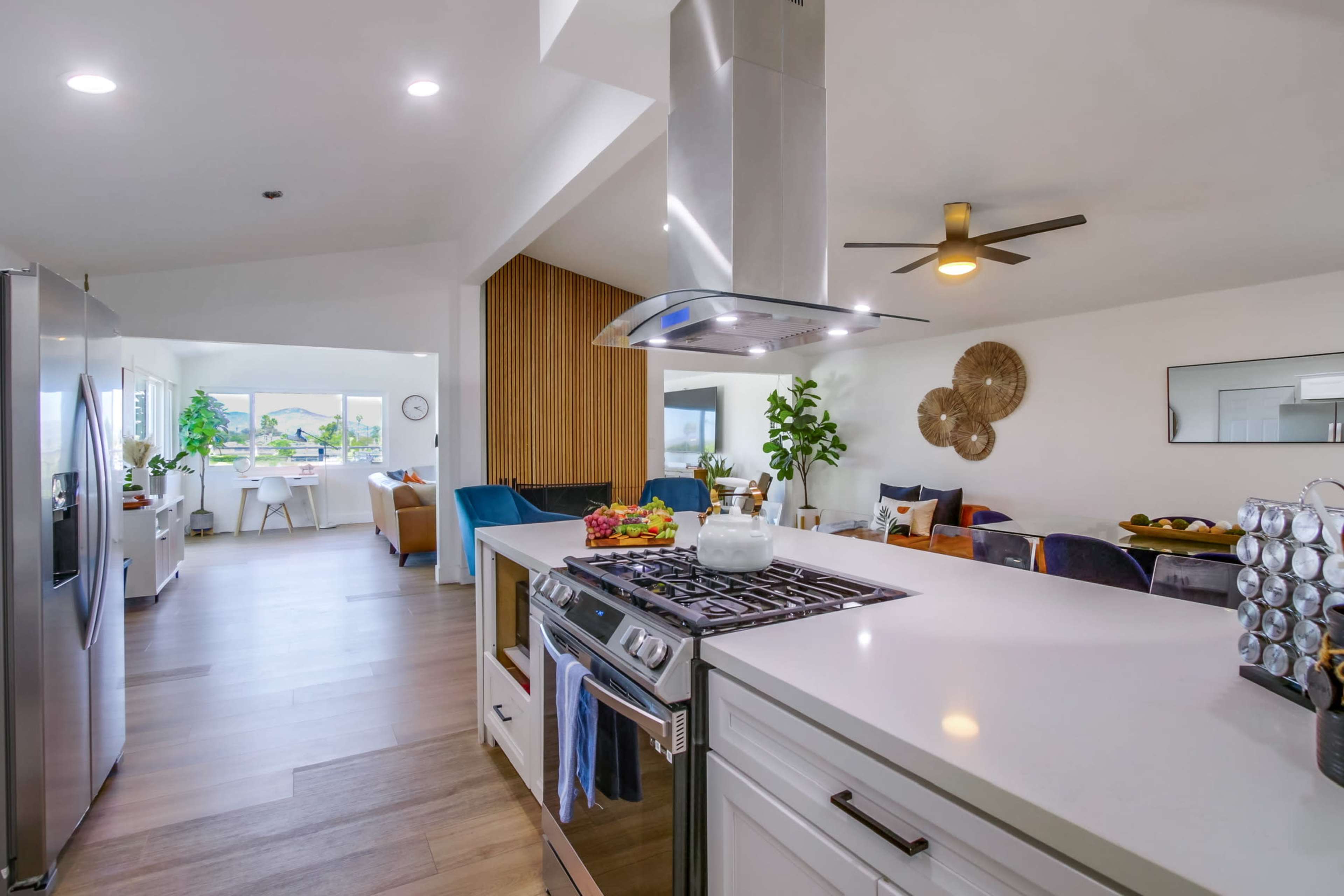 The image shows a modern kitchen with stainless steel appliances, a large island, and a living area visible in the background featuring seating and decorative elements.