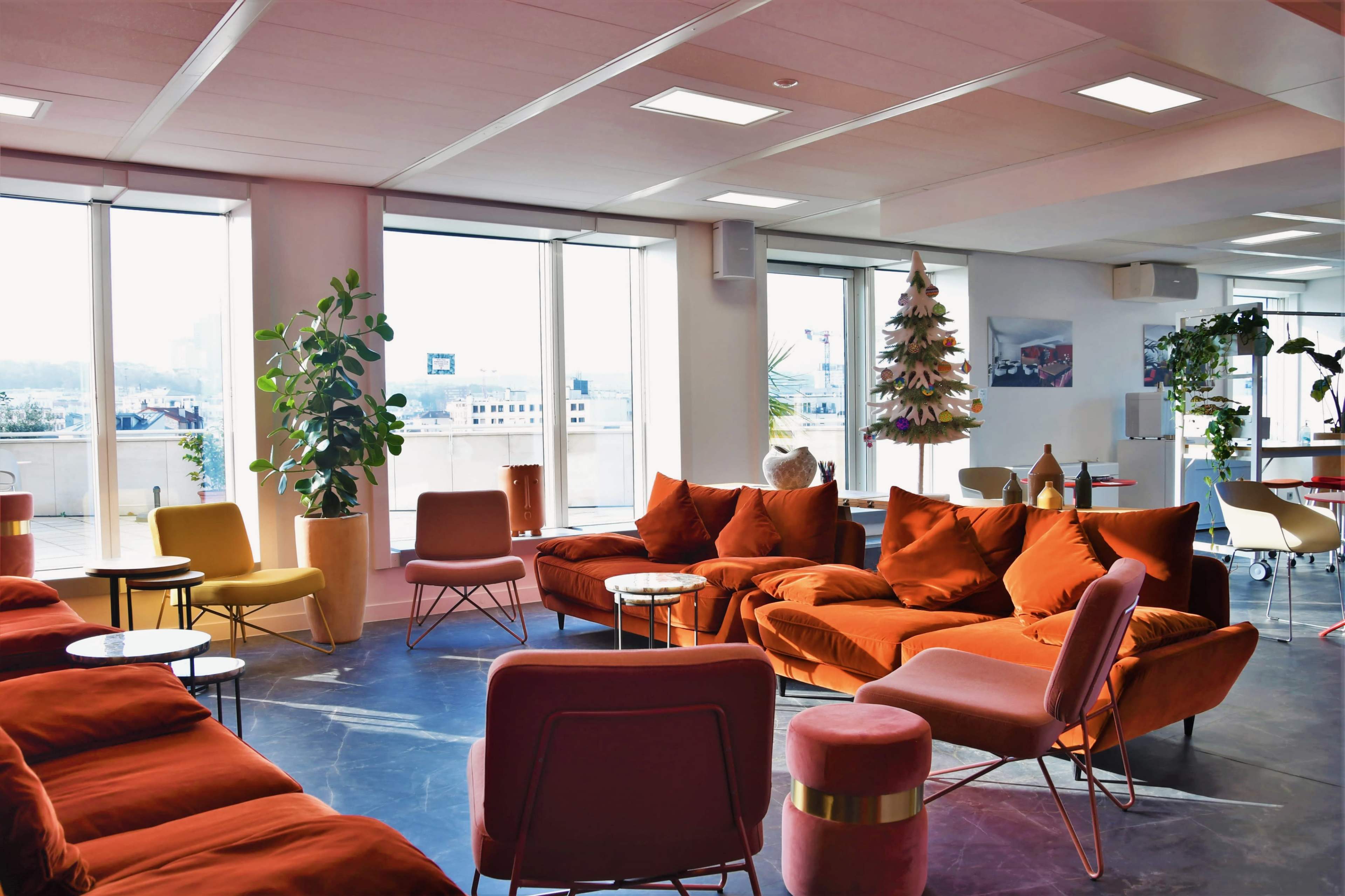 The image features a modern lounge area with orange and pink sofas, small round tables, and large windows overlooking the city.