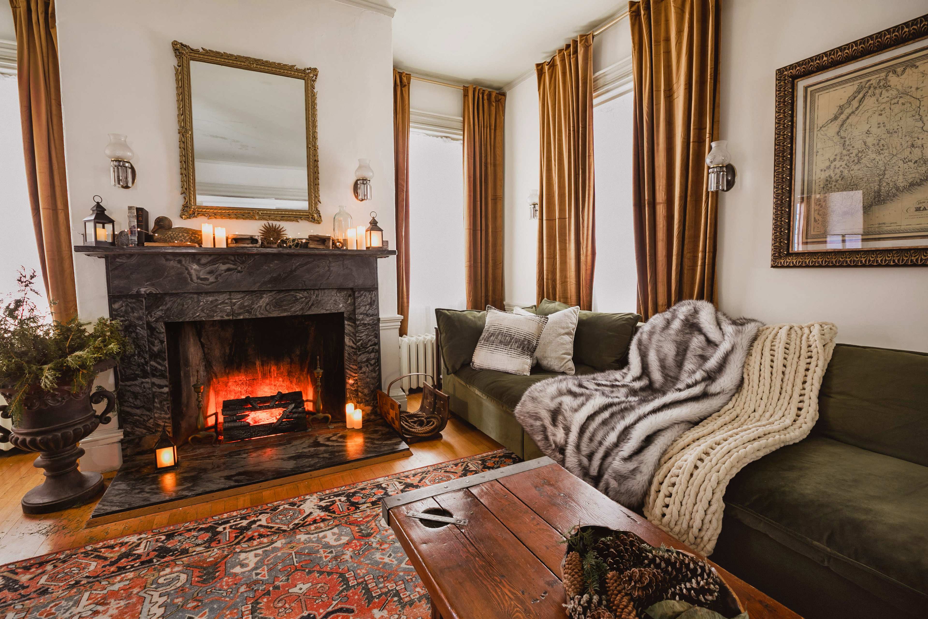A cozy living room features a dark marble fireplace, a plush green couch draped with a striped blanket, and a wooden coffee table, all illuminated by candles.