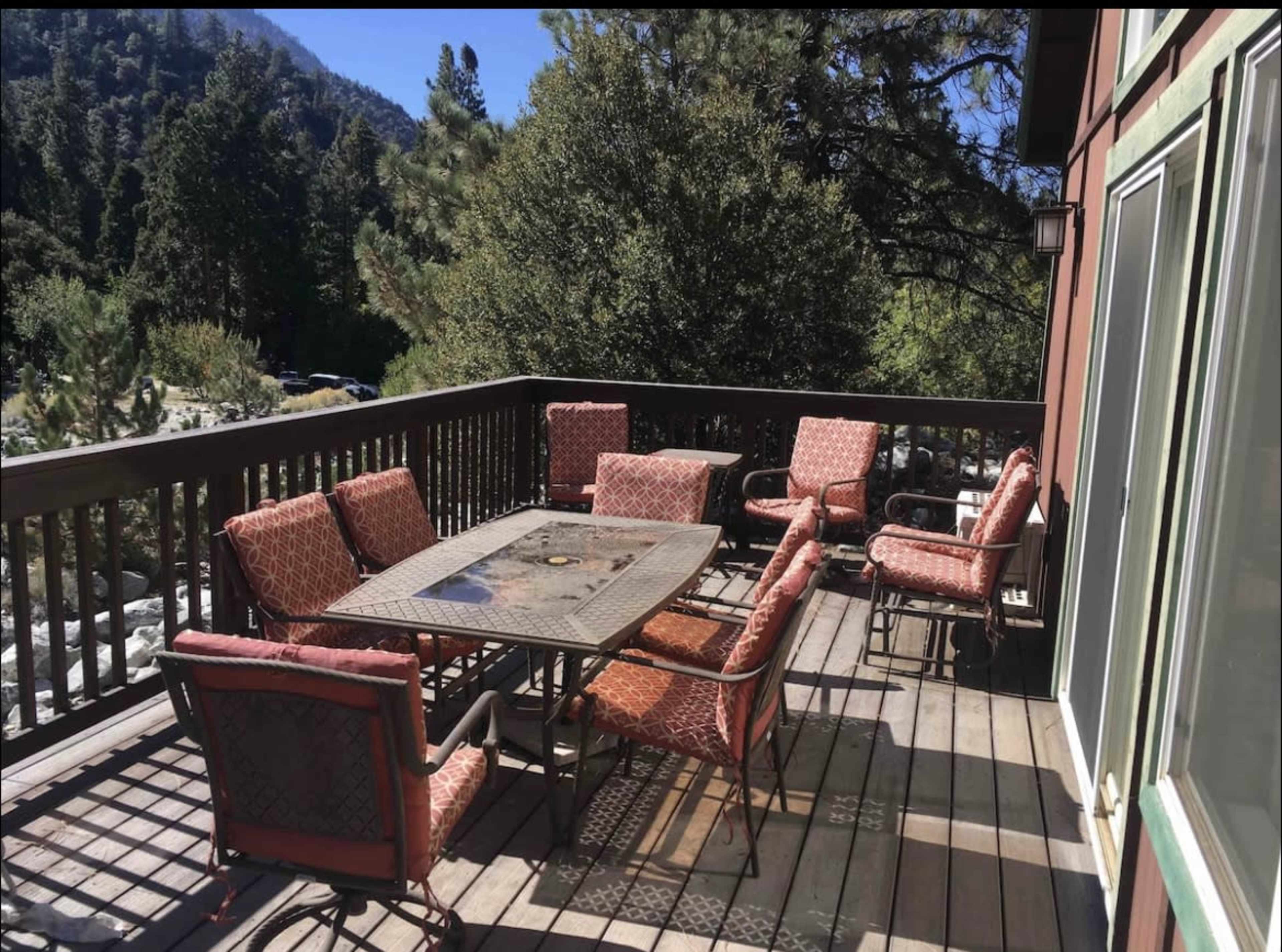 A wooden deck features a table surrounded by several cushioned chairs, set against a backdrop of trees and mountains.
