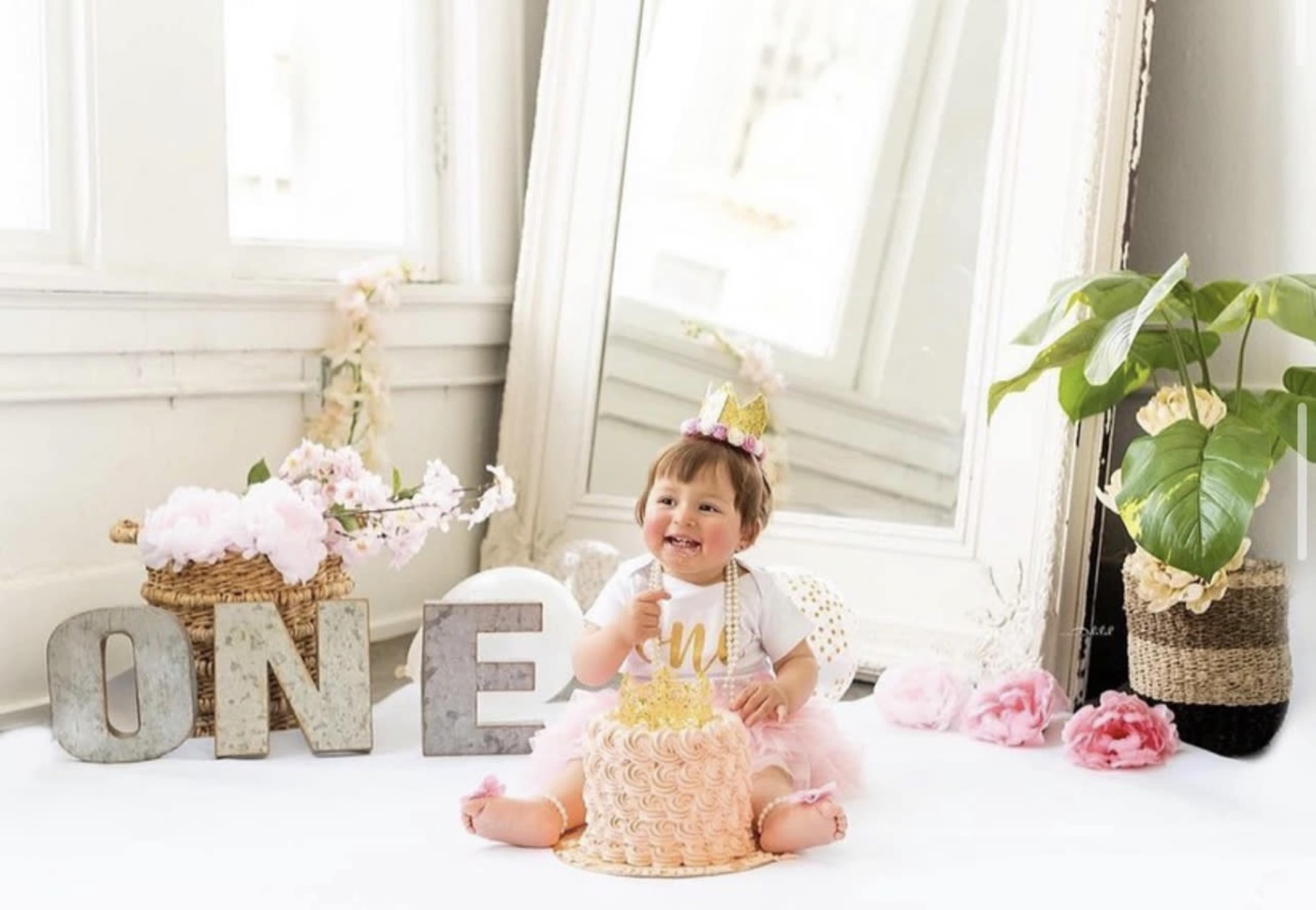 A child sits on a white surface next to a cake, surrounded by decorative flowers and the word "ONE" displayed in large letters.