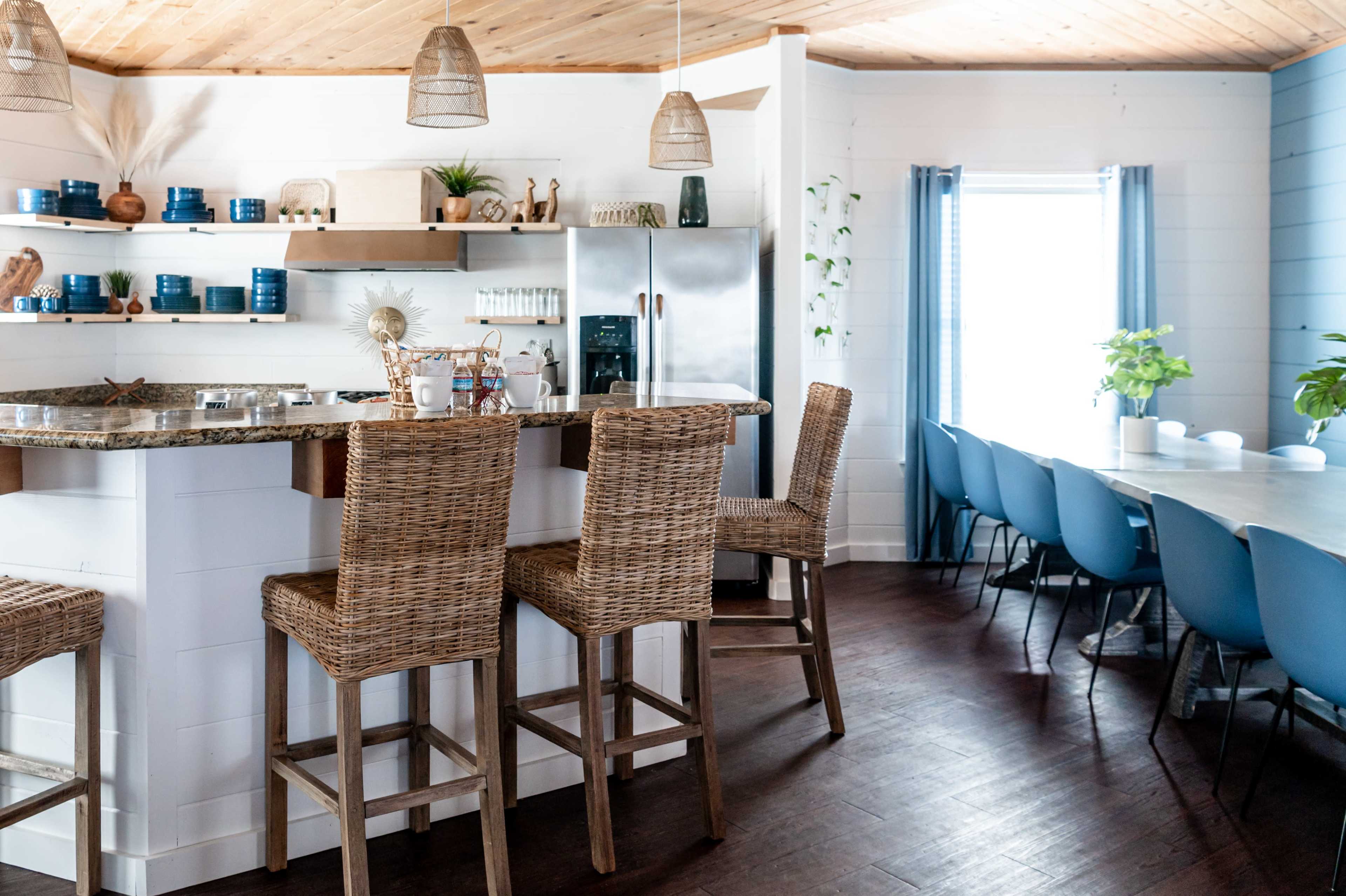 A modern kitchen features a bar with wicker stools and a dining table with blue chairs, complemented by light wood accents and plants.