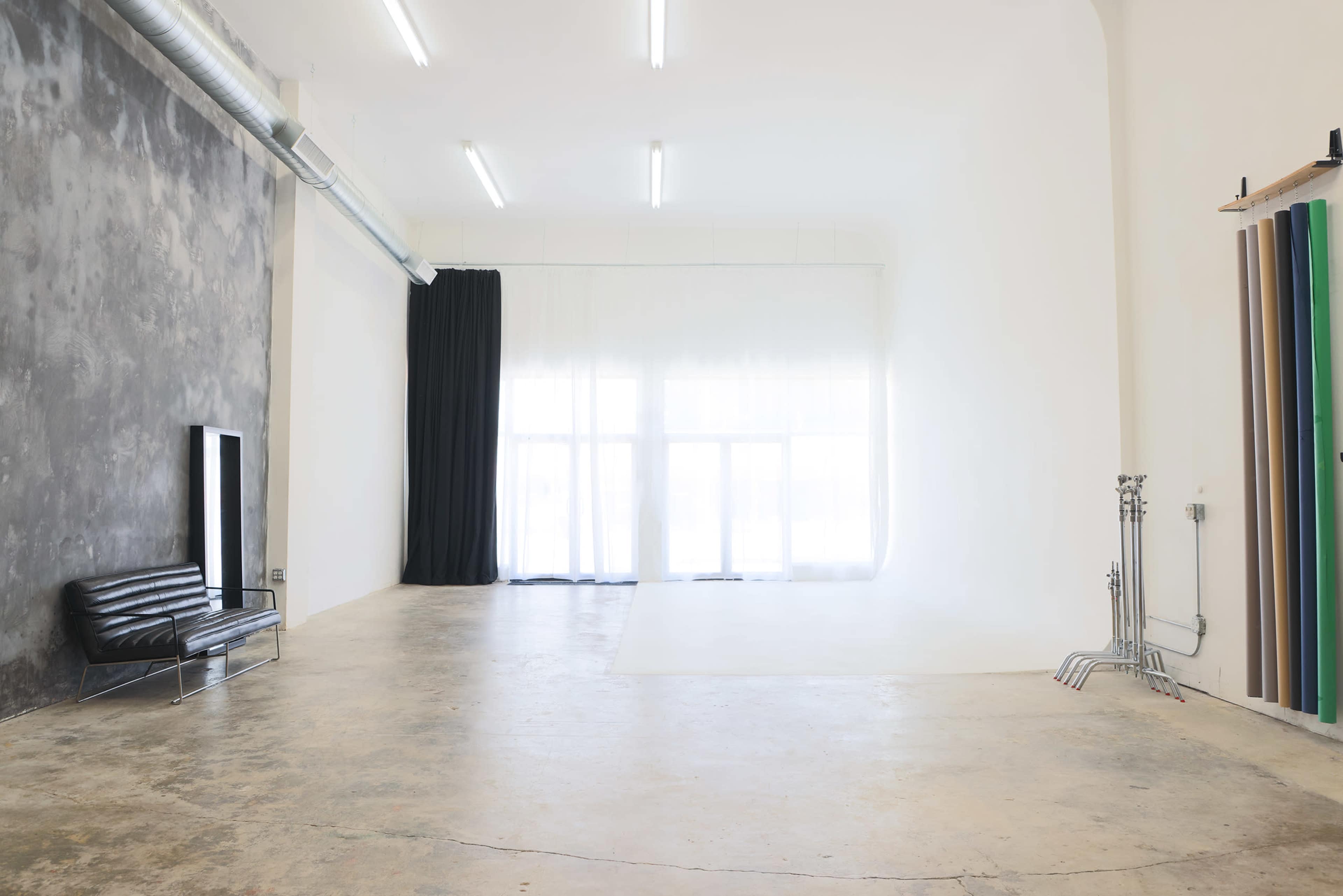 The image shows a spacious, empty studio with textured concrete and white walls, featuring a black couch and large windows covered by sheer curtains.
