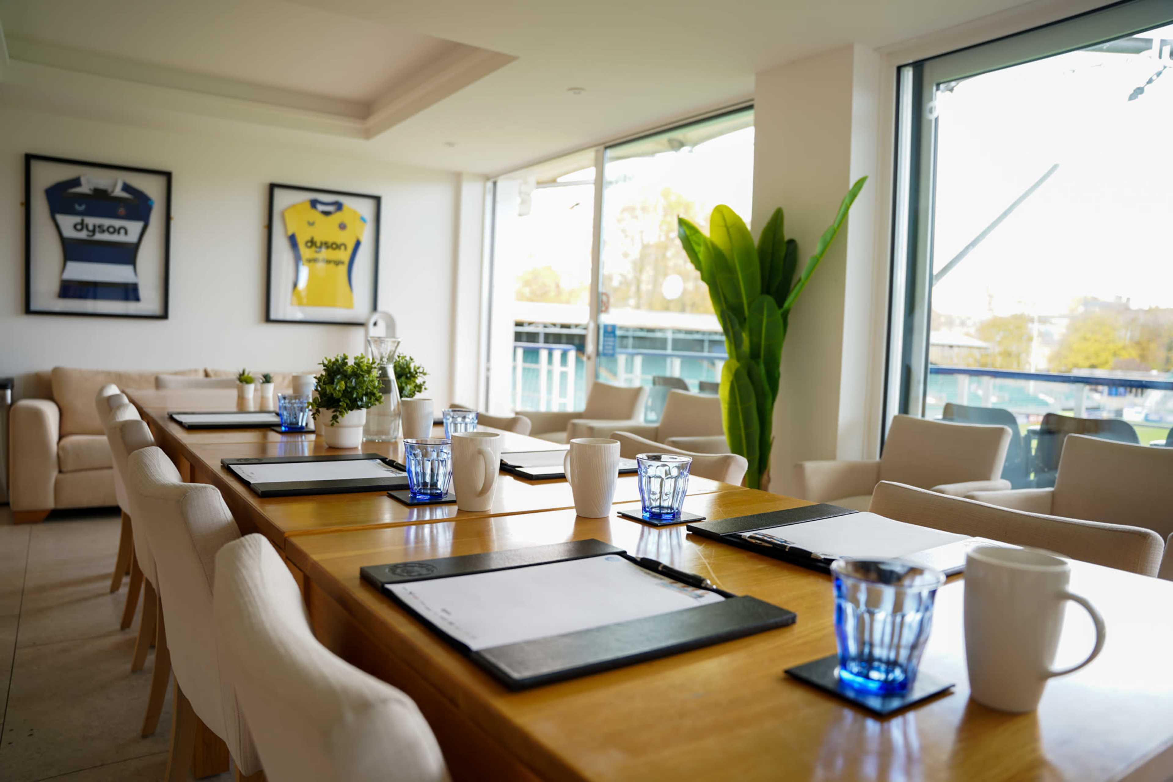 A modern meeting room features a long wooden table set with notepads and glasses, surrounded by comfortable chairs and framed jerseys on the walls.