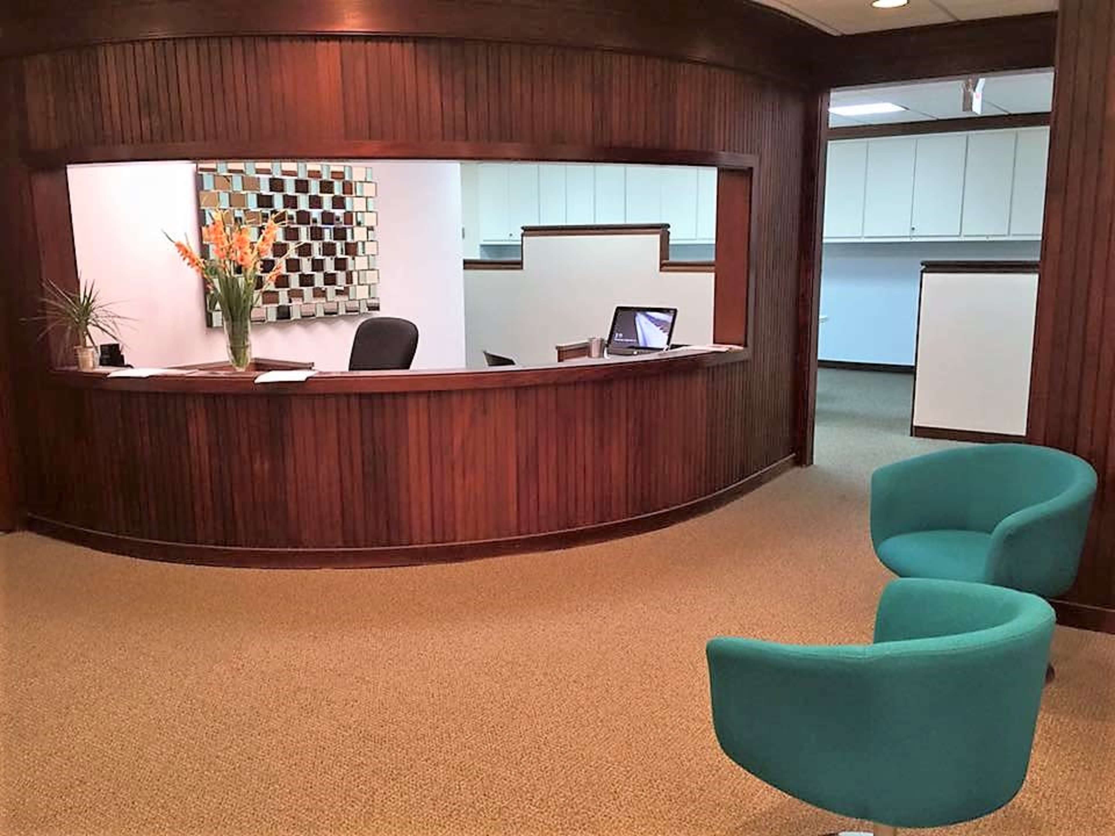 The image shows a reception area with a curved wooden desk, a computer, and two green chairs positioned in front of it.