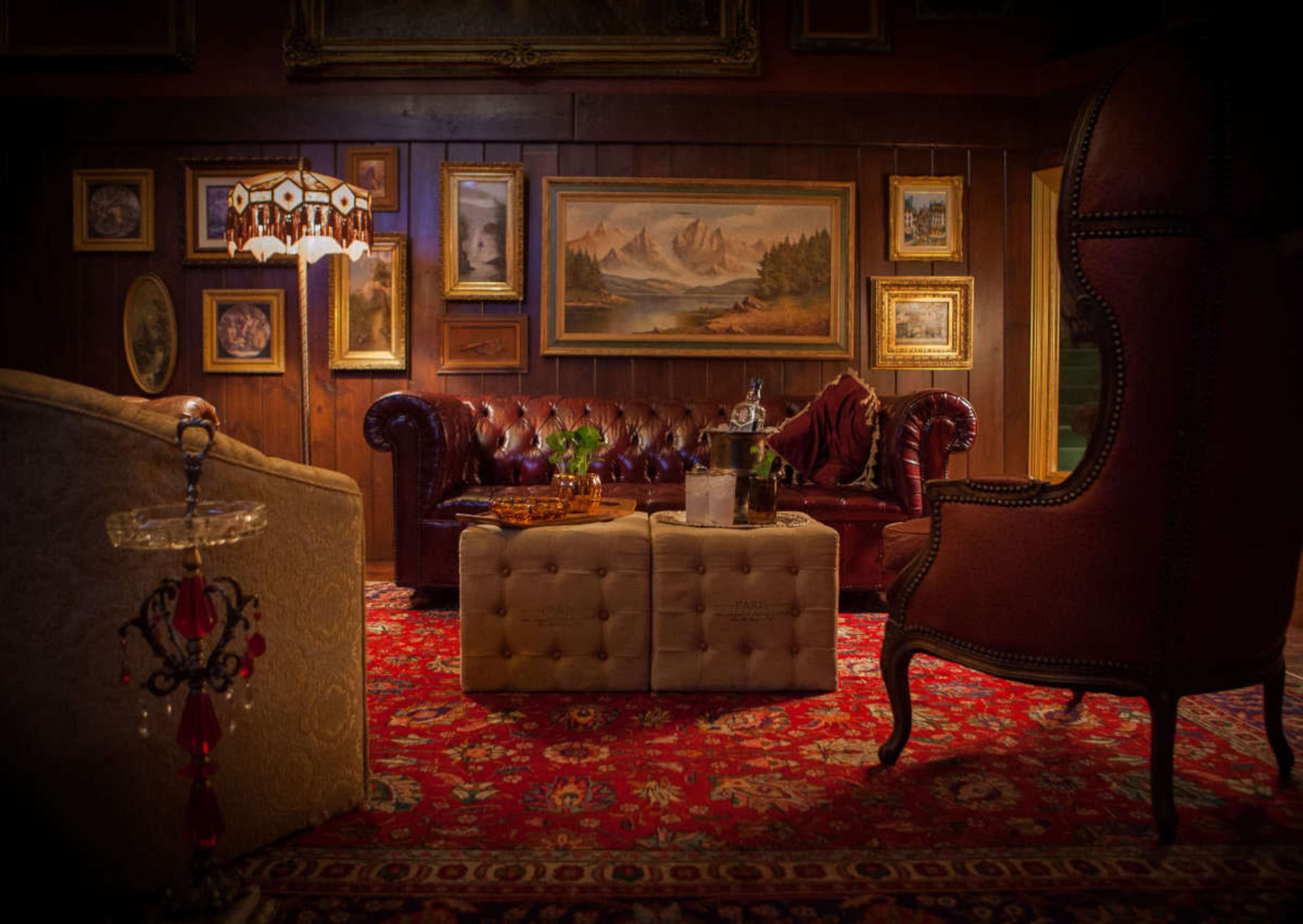 The image shows a cozy living room featuring a leather sofa, ornate armchair, a collection of framed artwork on the wooden walls, and a decorative area rug.