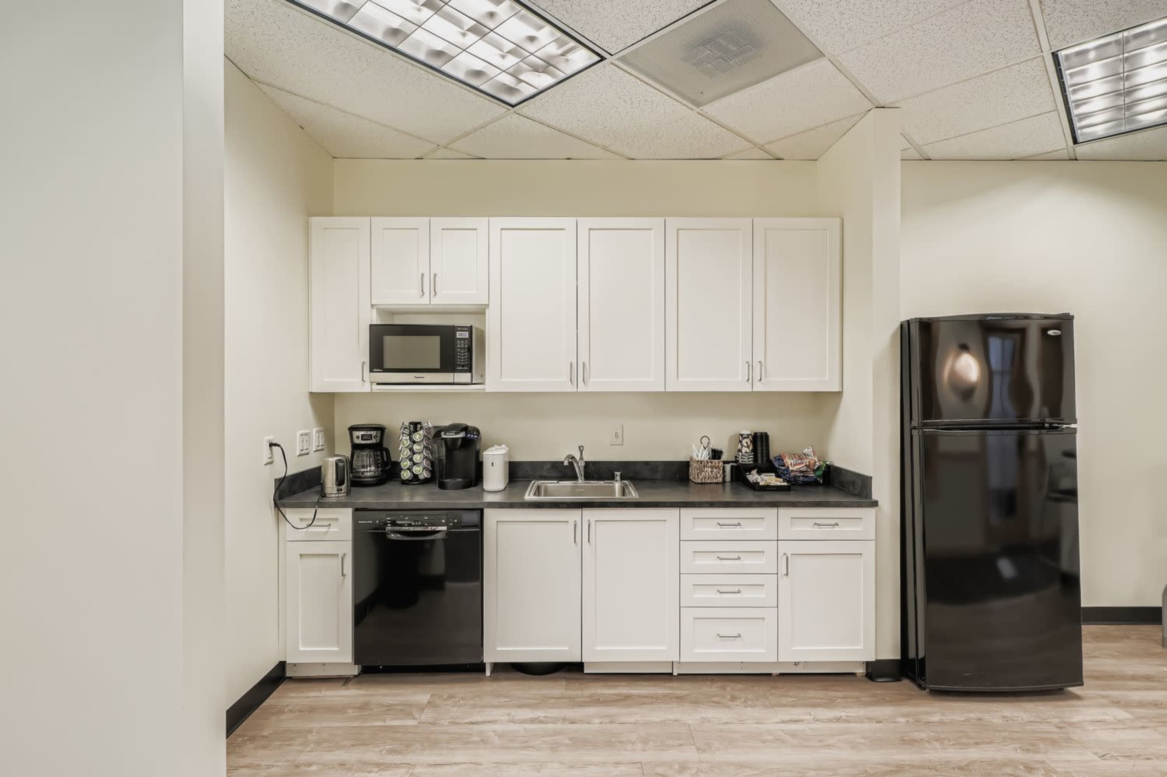 The image shows a modern kitchen space with white cabinets, a black countertop, and appliances including a microwave, coffee maker, and refrigerator.