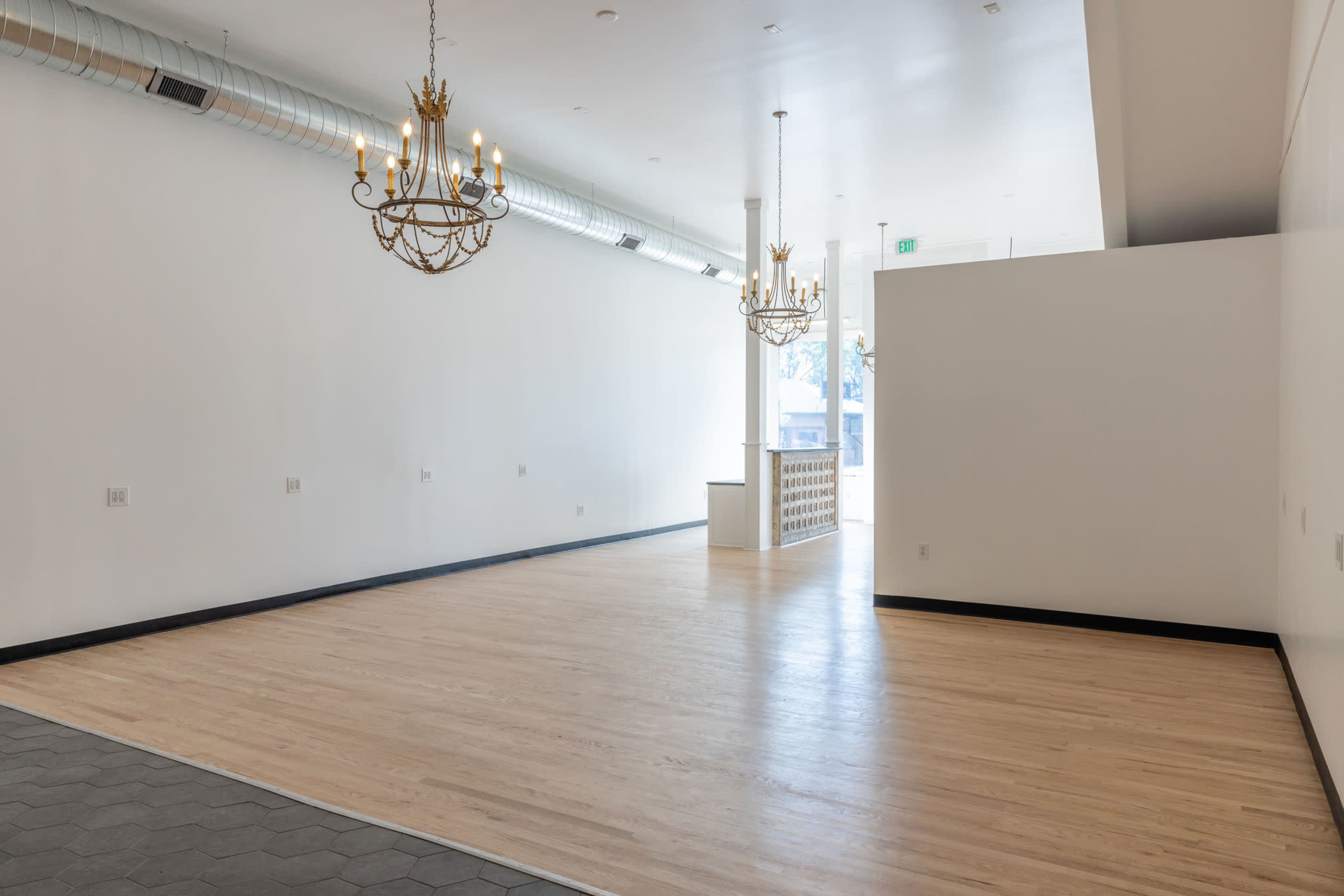 An empty room with wooden flooring, two chandeliers hanging from the ceiling, and light-colored walls, leading to a well-lit entrance.
