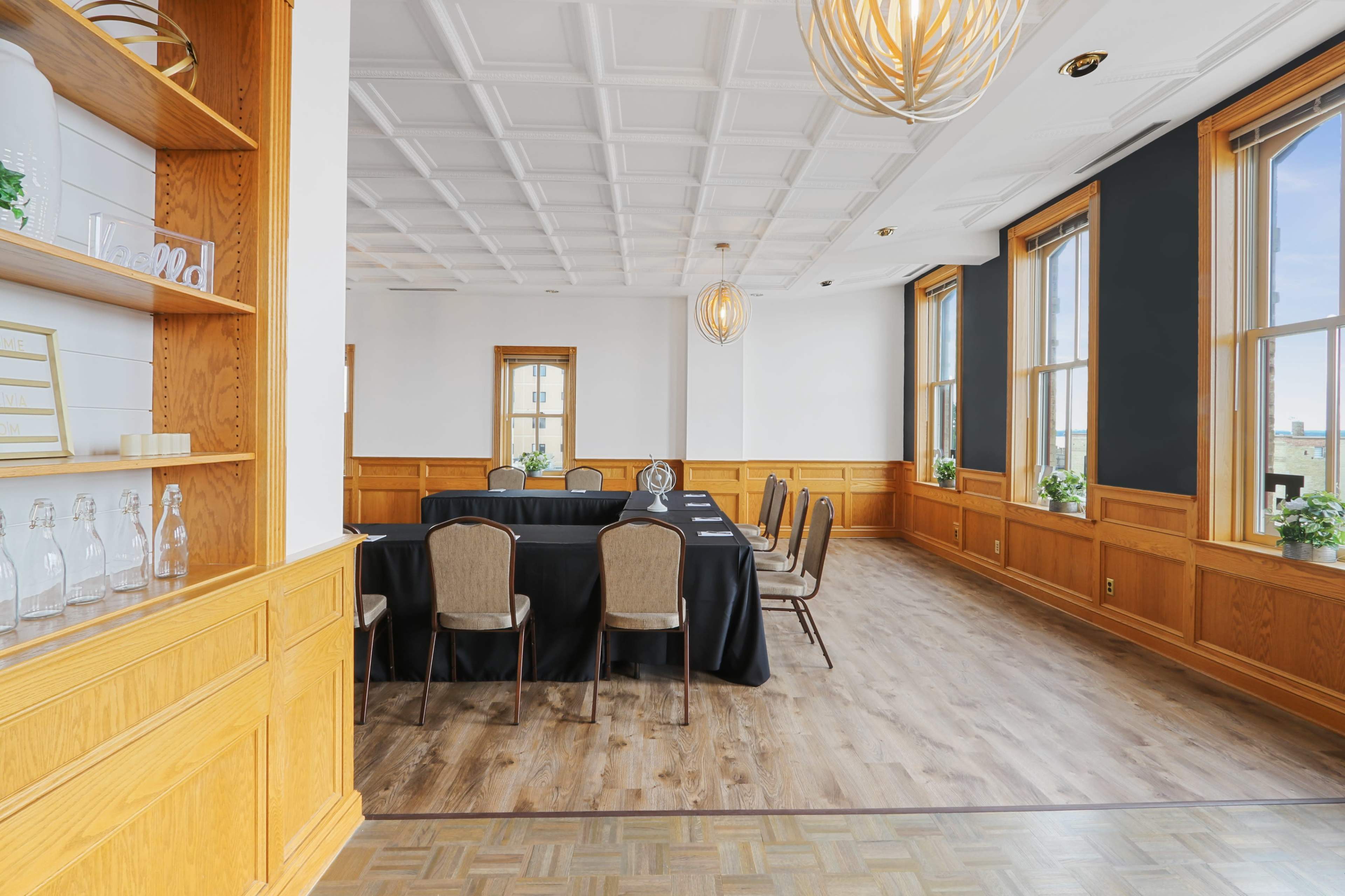 A conference room with a long black table surrounded by chairs, set against large windows with wooden frames and decorative light fixtures.