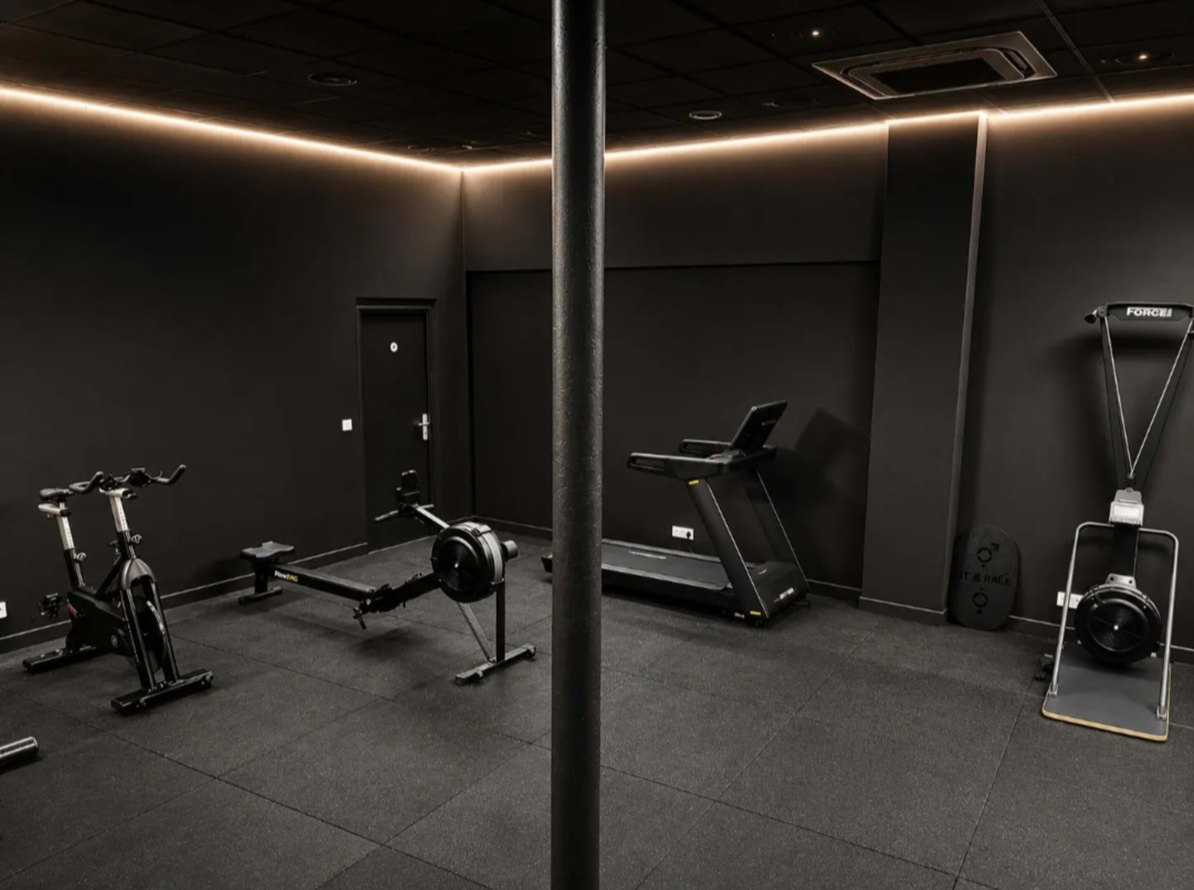 The image shows a dark-themed gym featuring cardio equipment including a treadmill and rowing machine, along with exercise bikes and a central support column.