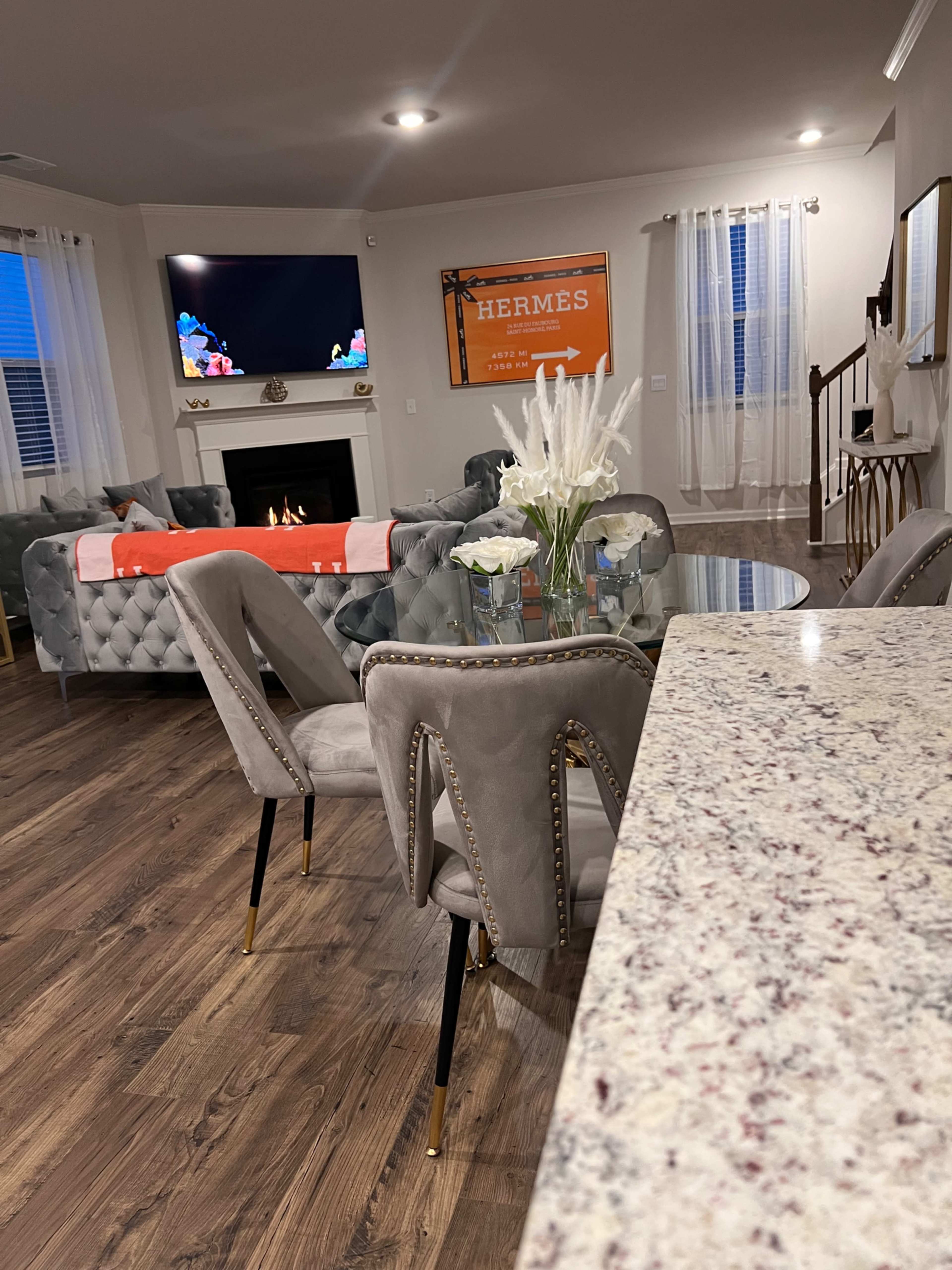 A modern living room features a glass dining table surrounded by upholstered chairs, with a TV mounted above a fireplace and orange wall art in the background.