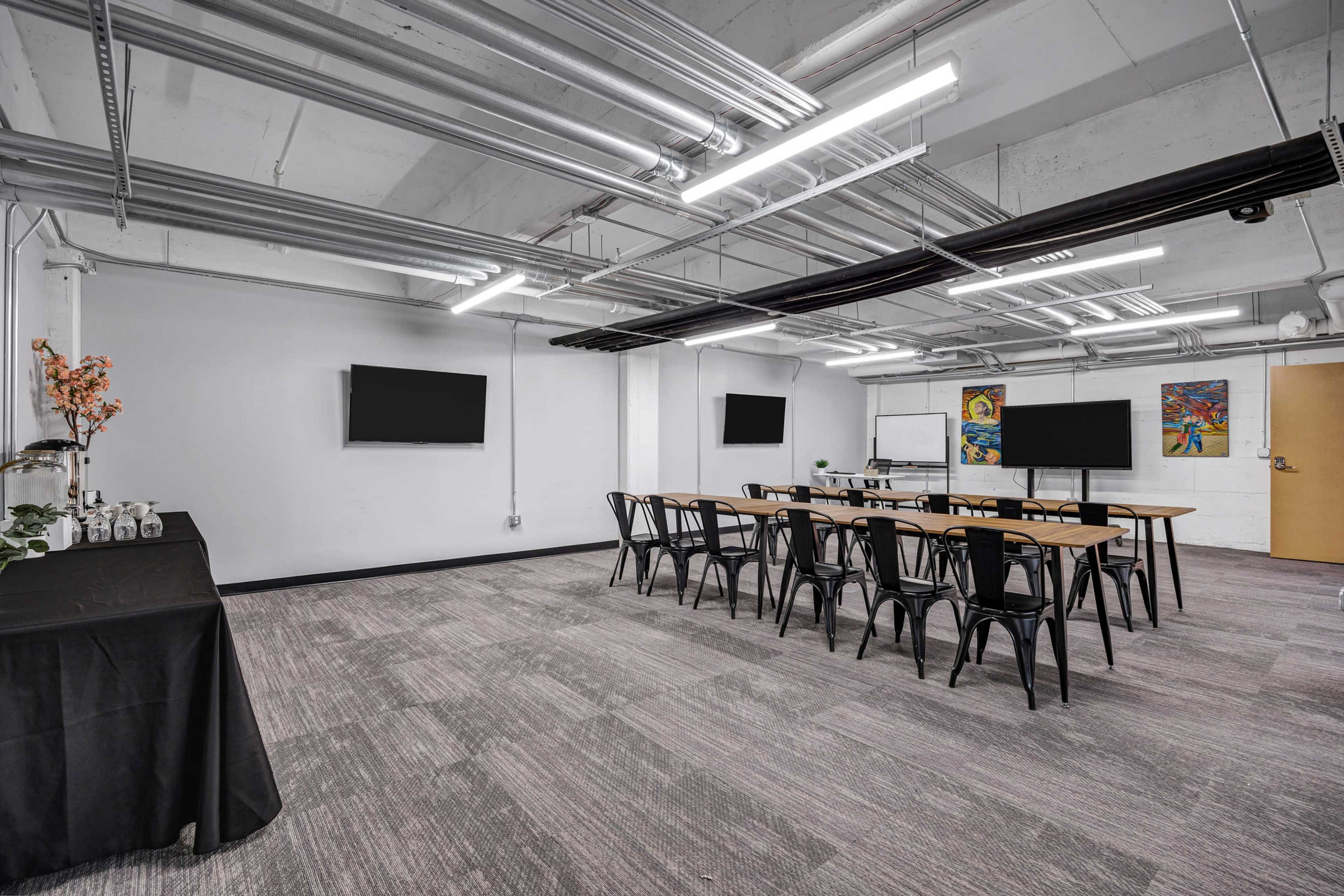 The image shows a modern meeting room with two large screens, a long table surrounded by chairs, and decorative elements on a side table.