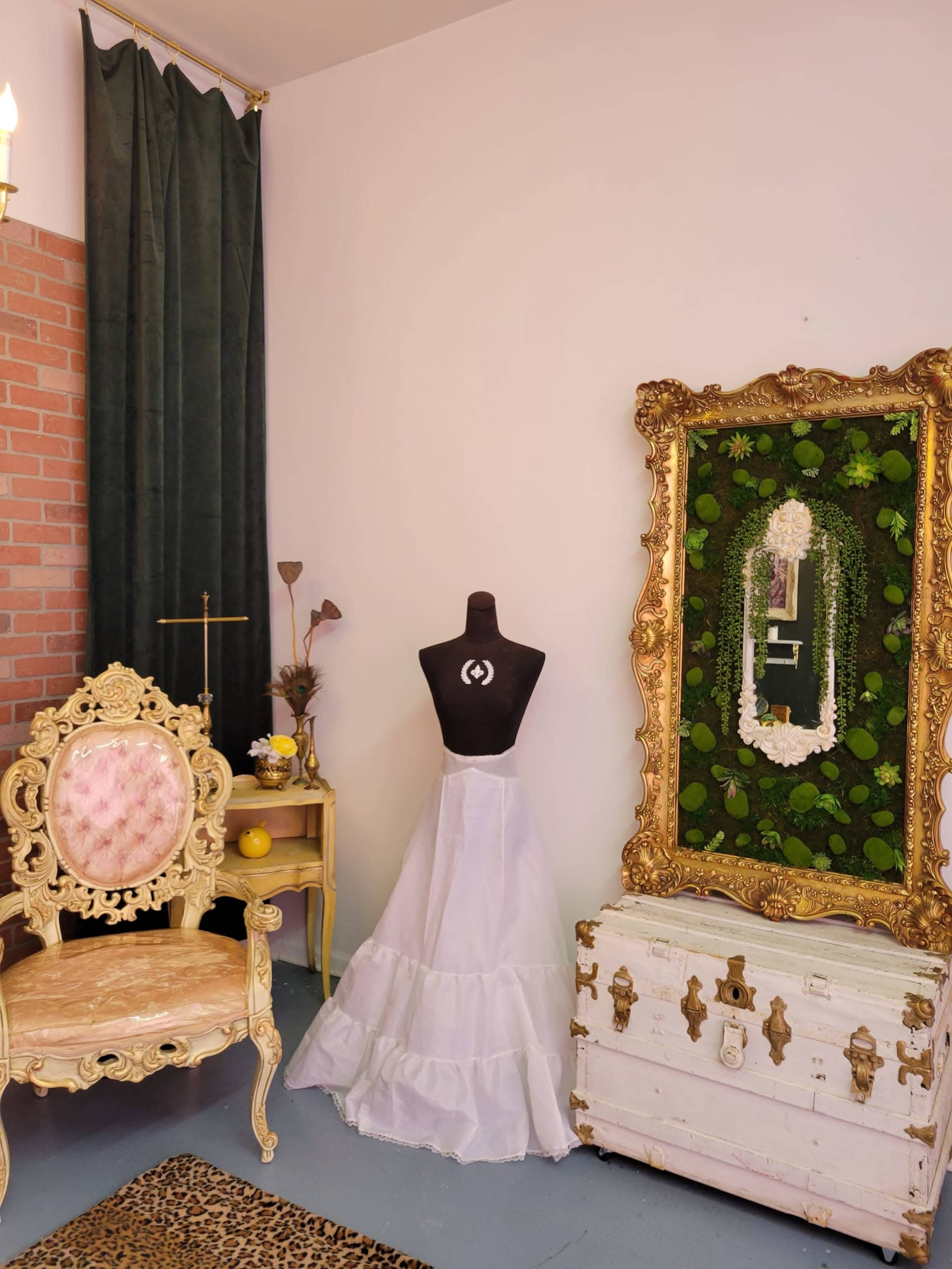 A vintage-inspired corner with a dress form displaying a white gown, a decorative gold-framed mirror covered in greenery, a wooden chest, and an ornate chair.