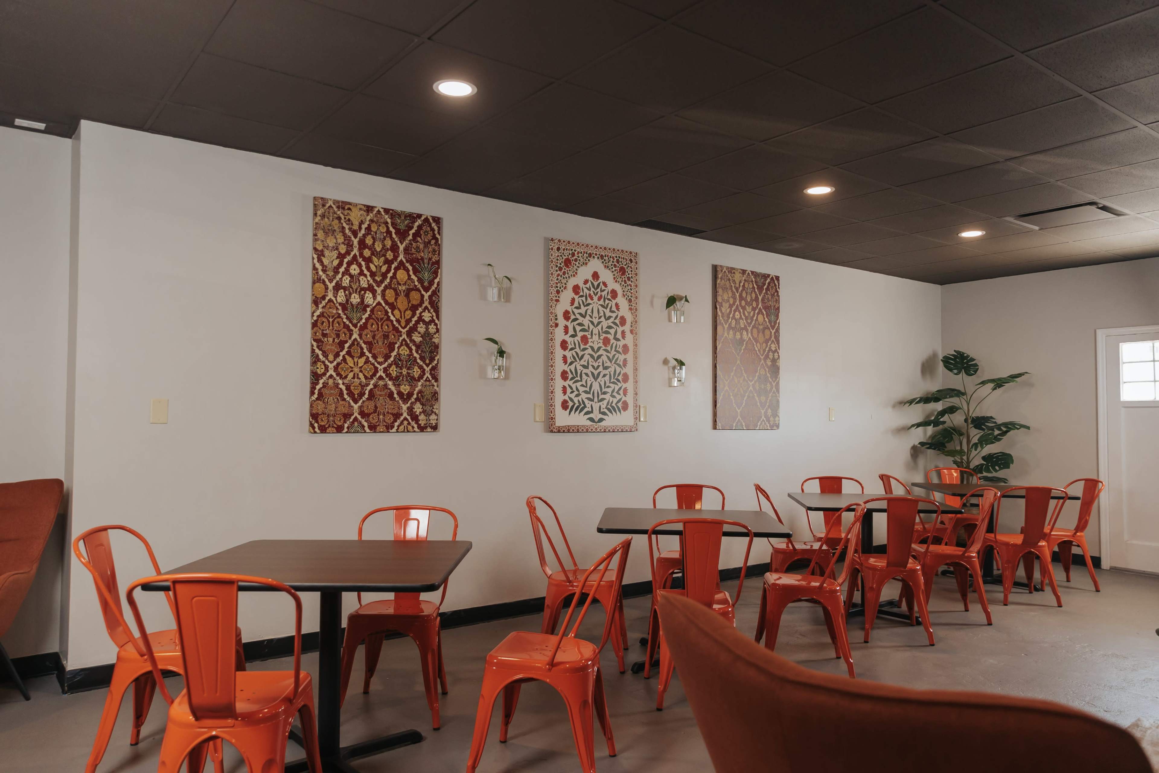 A cafe interior with orange metal chairs and tables, decorated walls featuring patterned artworks, and a few plants.