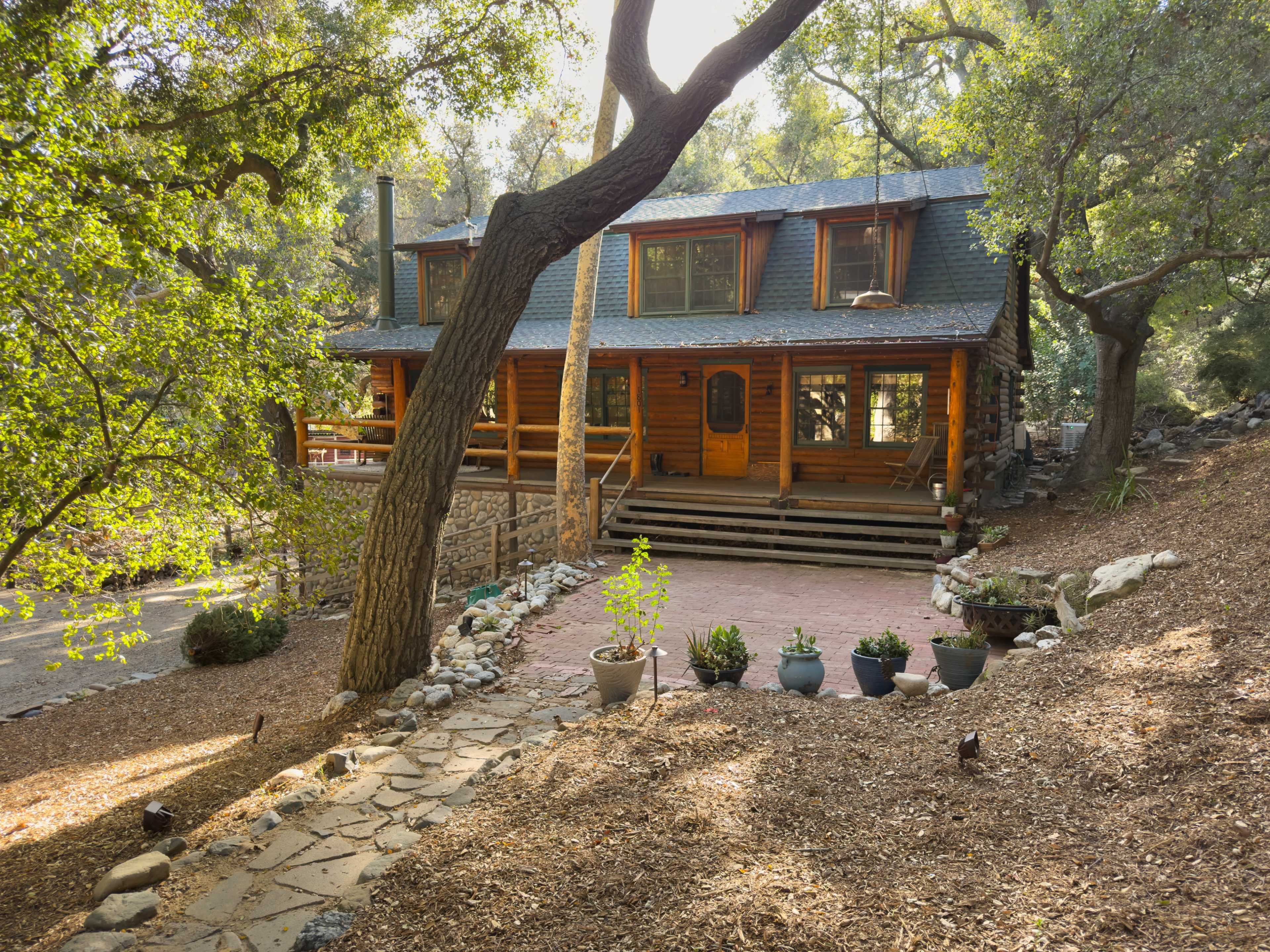 A log cabin situated in a wooded area features multiple windows and a front porch, surrounded by decorative stone pathways and potted plants.