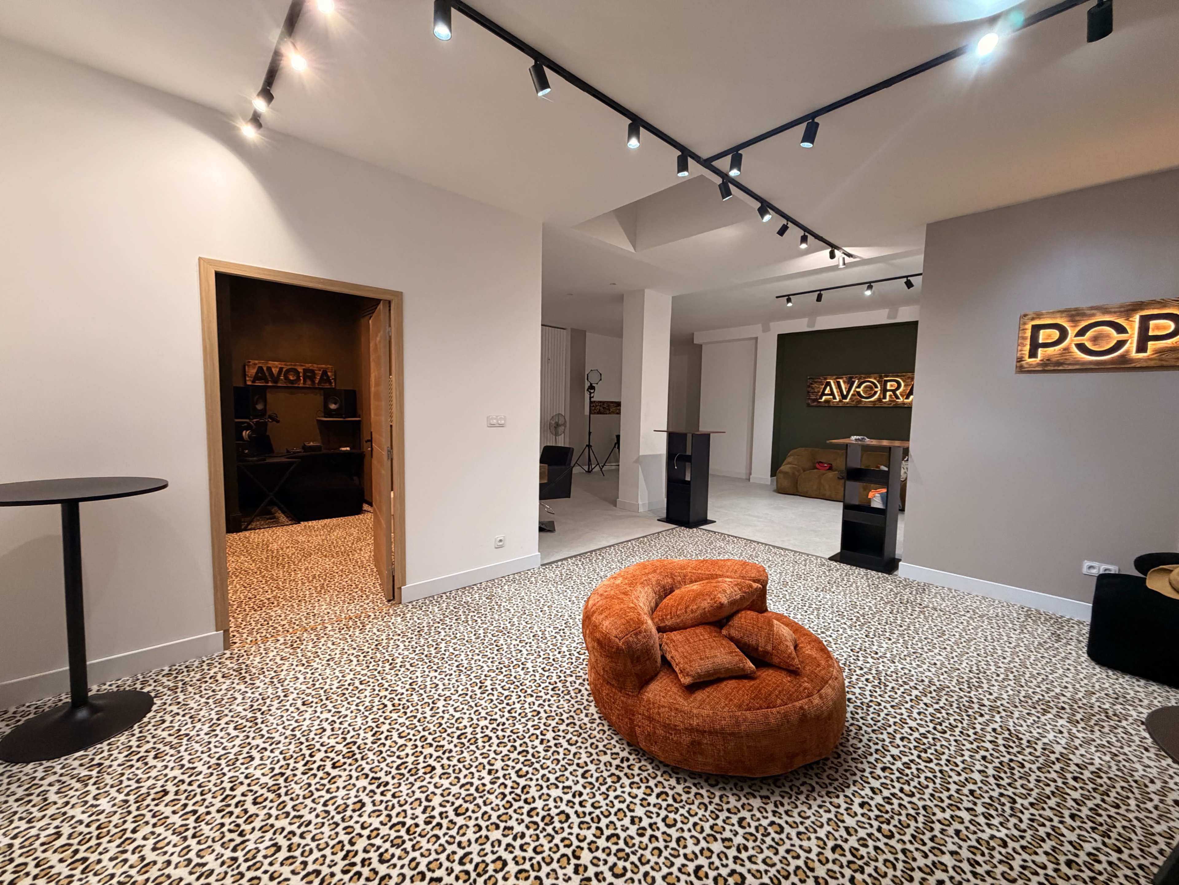 The image shows a modern interior space featuring a leopard-print carpet, an orange lounge chair, and illuminated signs labeled "AVORA" and "POP."