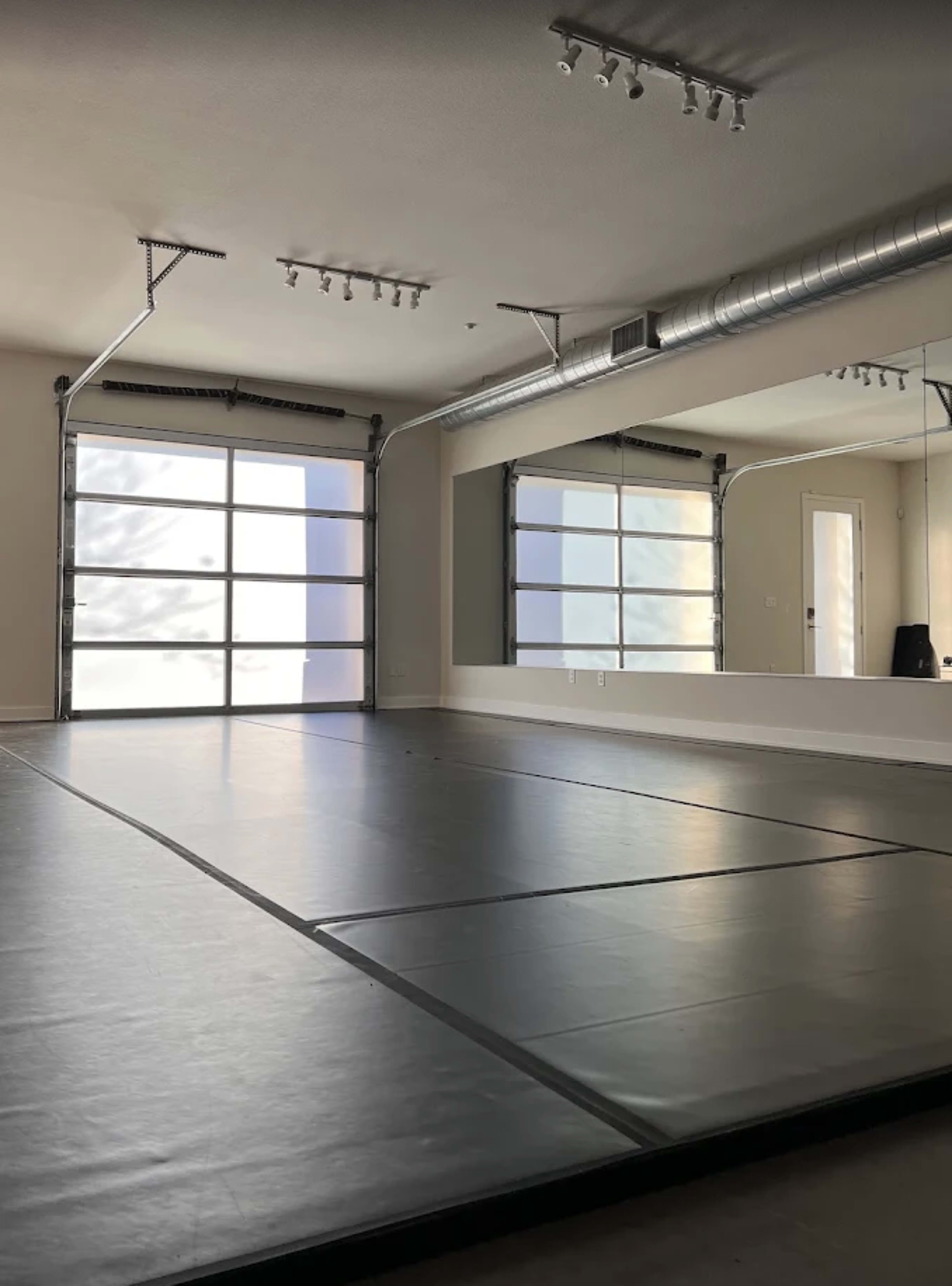 The image shows a spacious, empty room with a mirrored wall and a large garage-style door allowing natural light to enter.