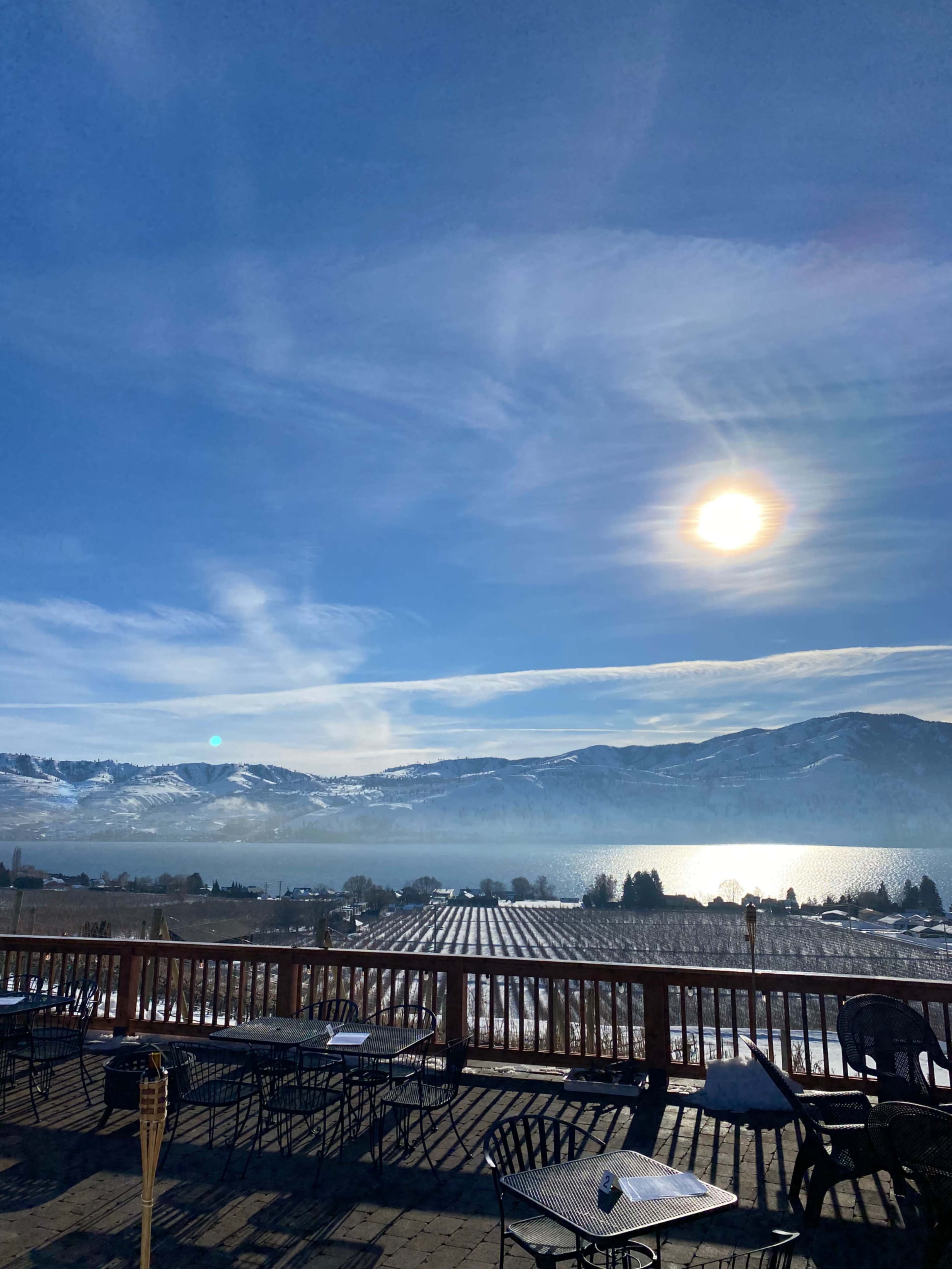 A snowy landscape with vineyards and a lake, framed by mountains under a clear blue sky with a bright sun.