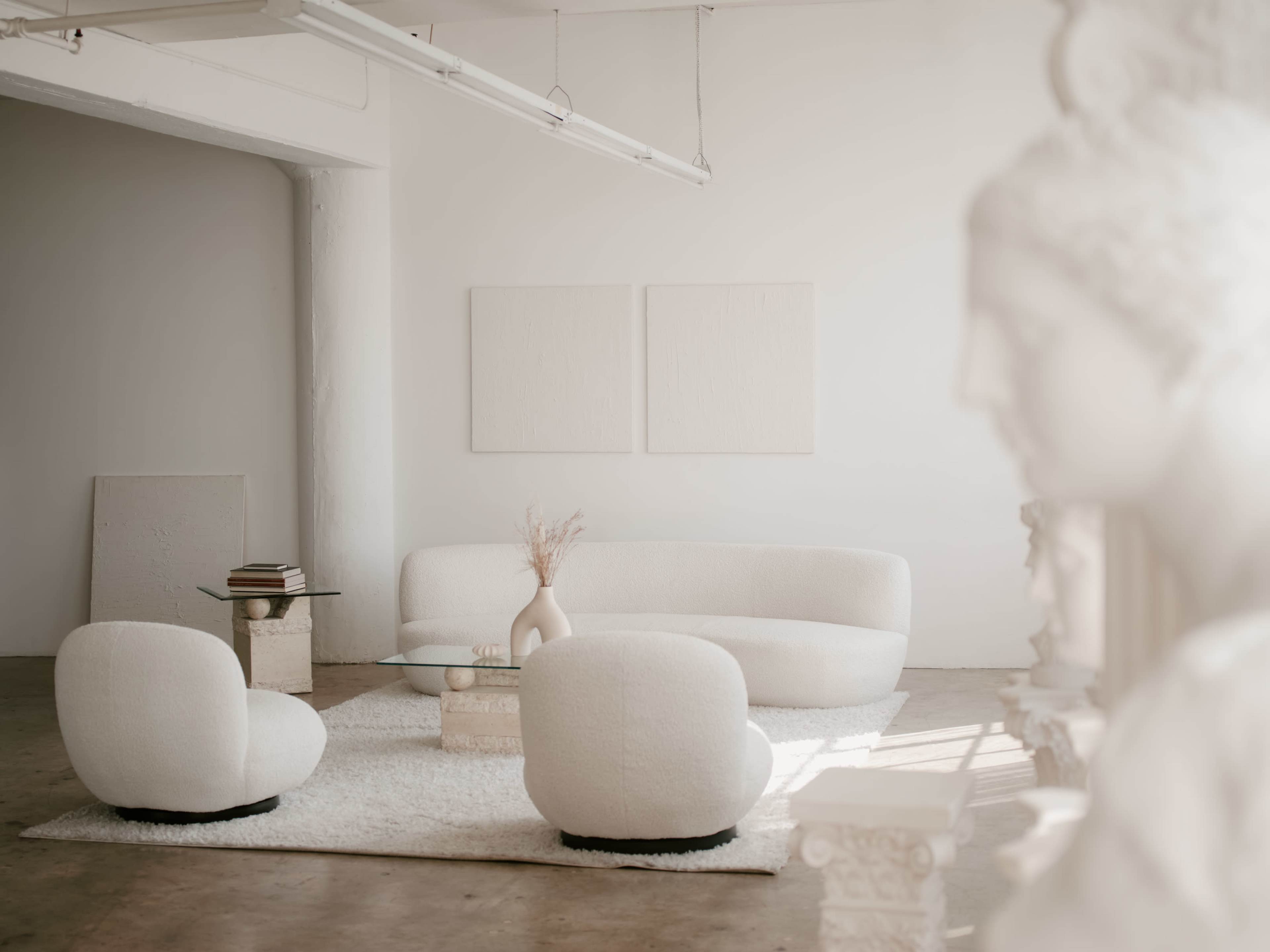 The scene depicts a minimalist, light-filled interior with a curved white couch, matching chairs, a glass coffee table, and a sculpture partially visible on the right.