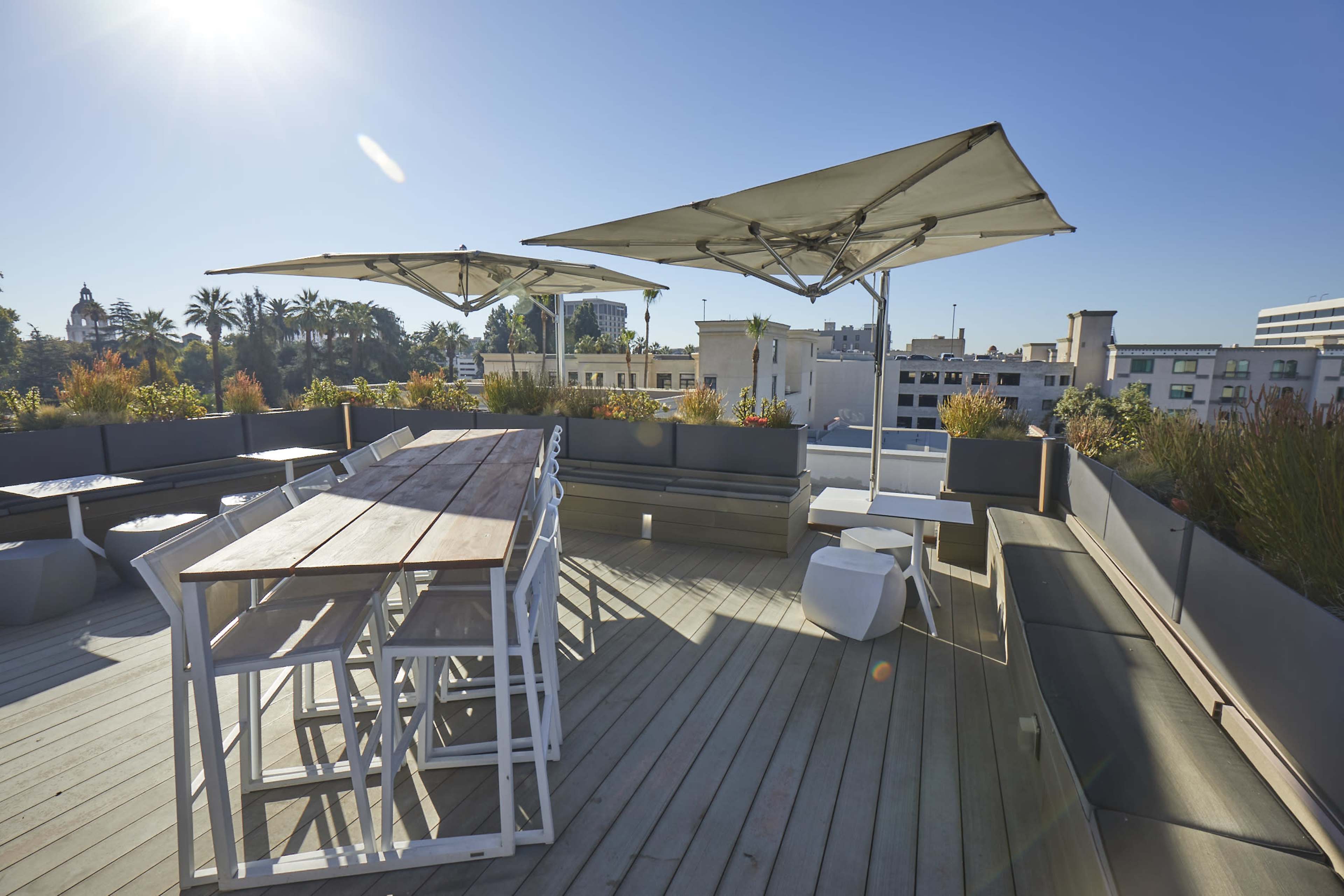 A rooftop terrace with large umbrellas, a wooden table, and benches, surrounded by potted plants against a clear blue sky.