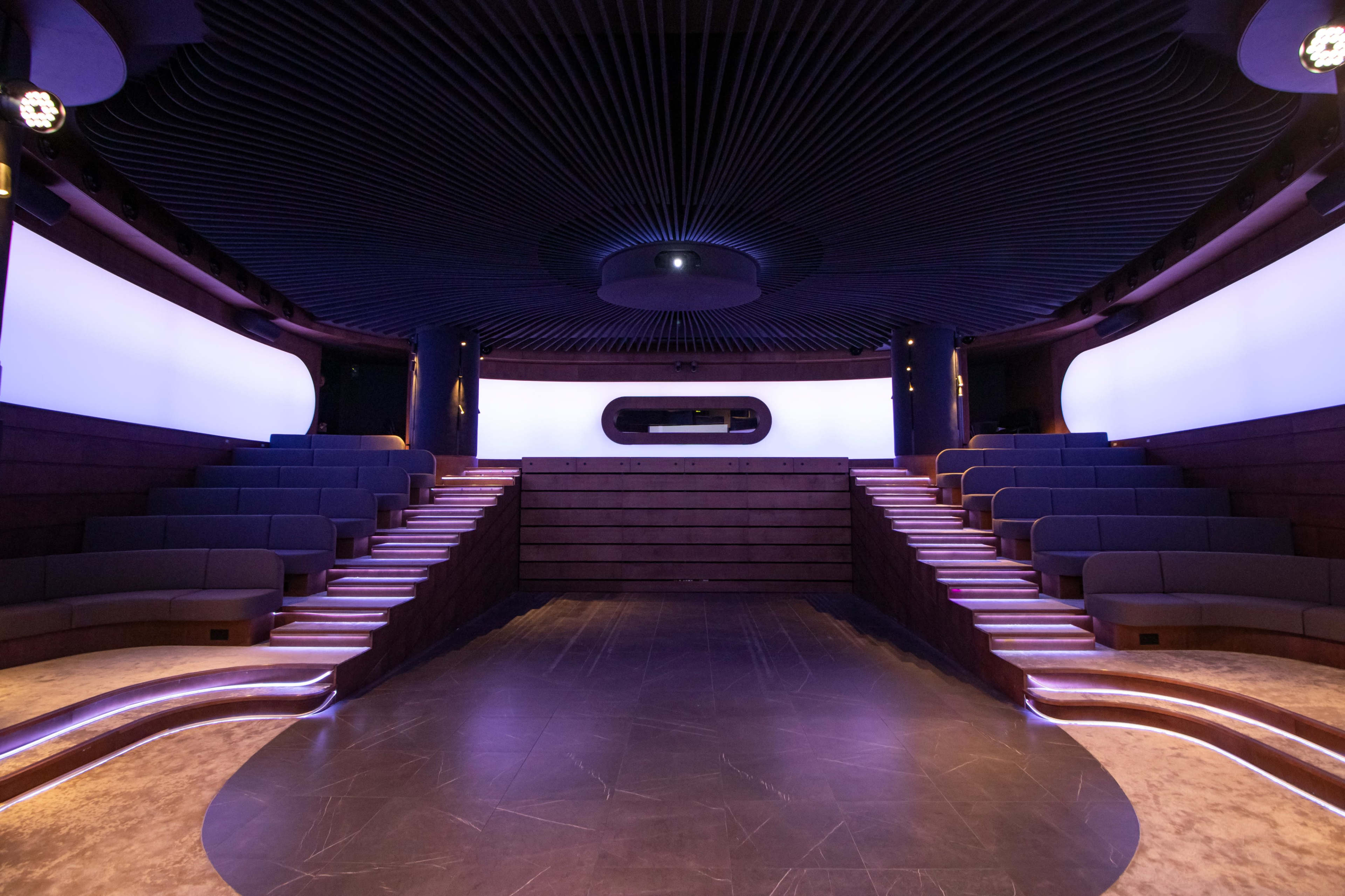 The image shows a modern, tiered auditorium with dark seating and curved, illuminated walls.