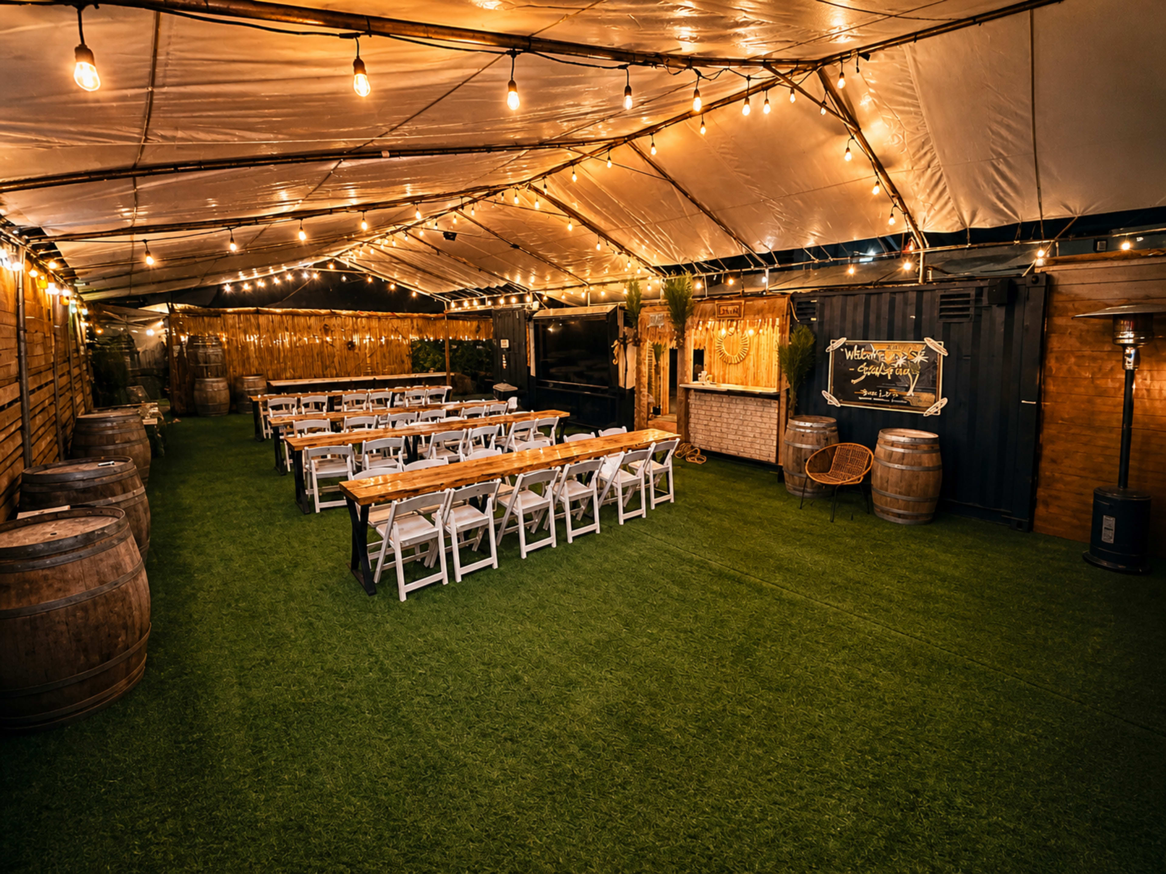 A large tented outdoor space is set up for an event, featuring rows of tables and chairs, string lights overhead, and decorative elements like barrels and greenery.