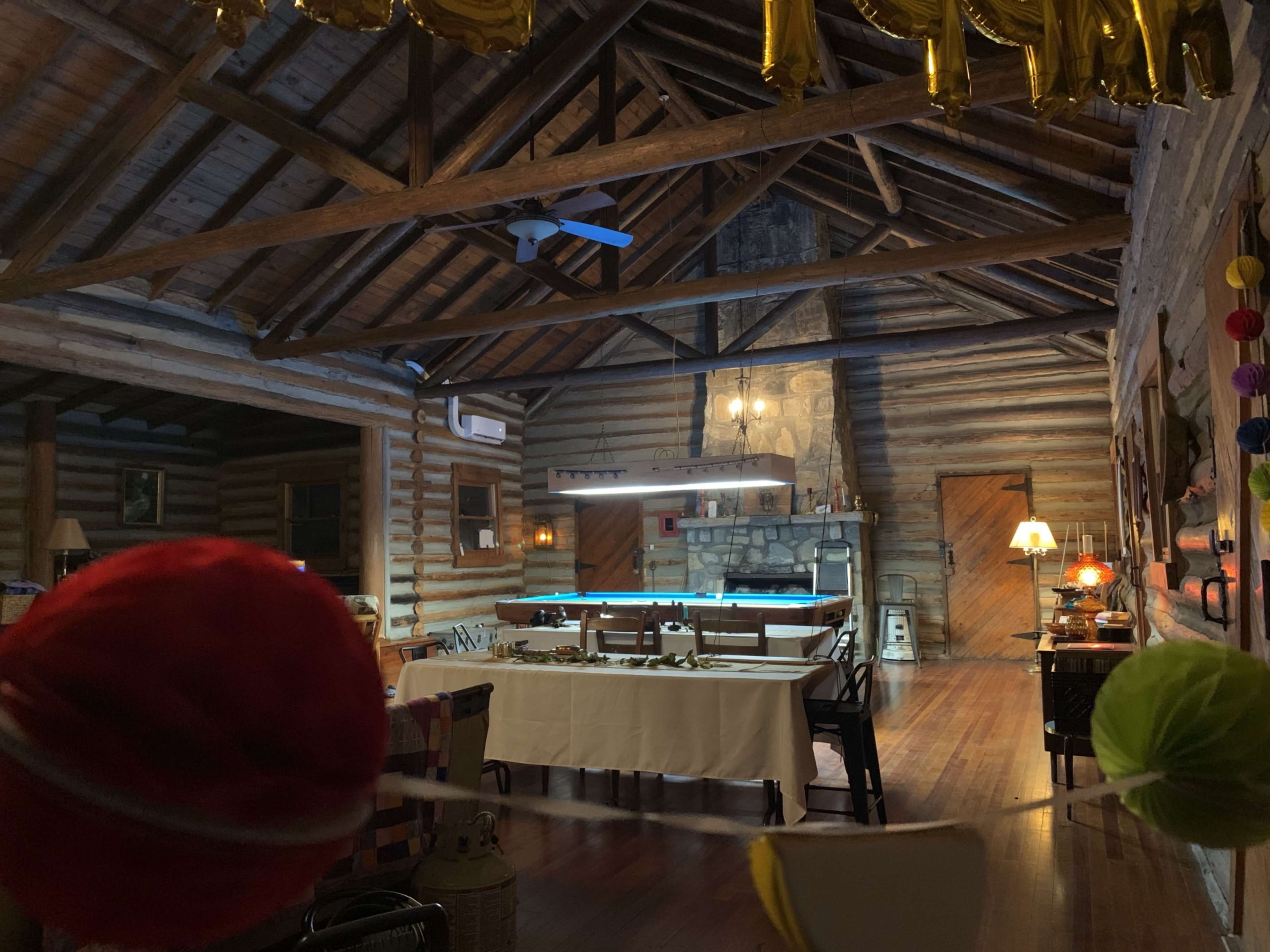 A spacious wooden interior of a rustic cabin, featuring exposed beams, a dining area with tables, and a stone fireplace.