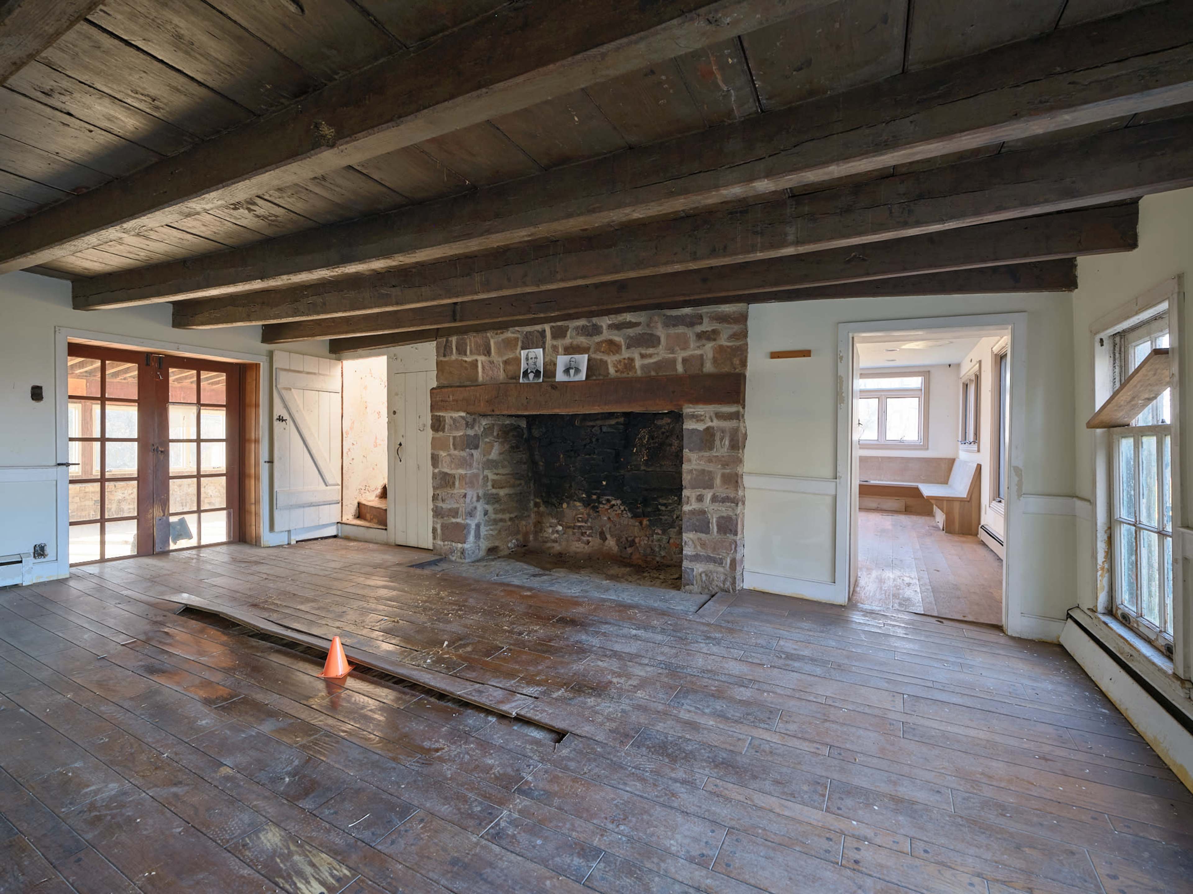 An empty room with wooden beams and a stone fireplace at one end, featuring exposed floorboards and a small orange traffic cone in the center.