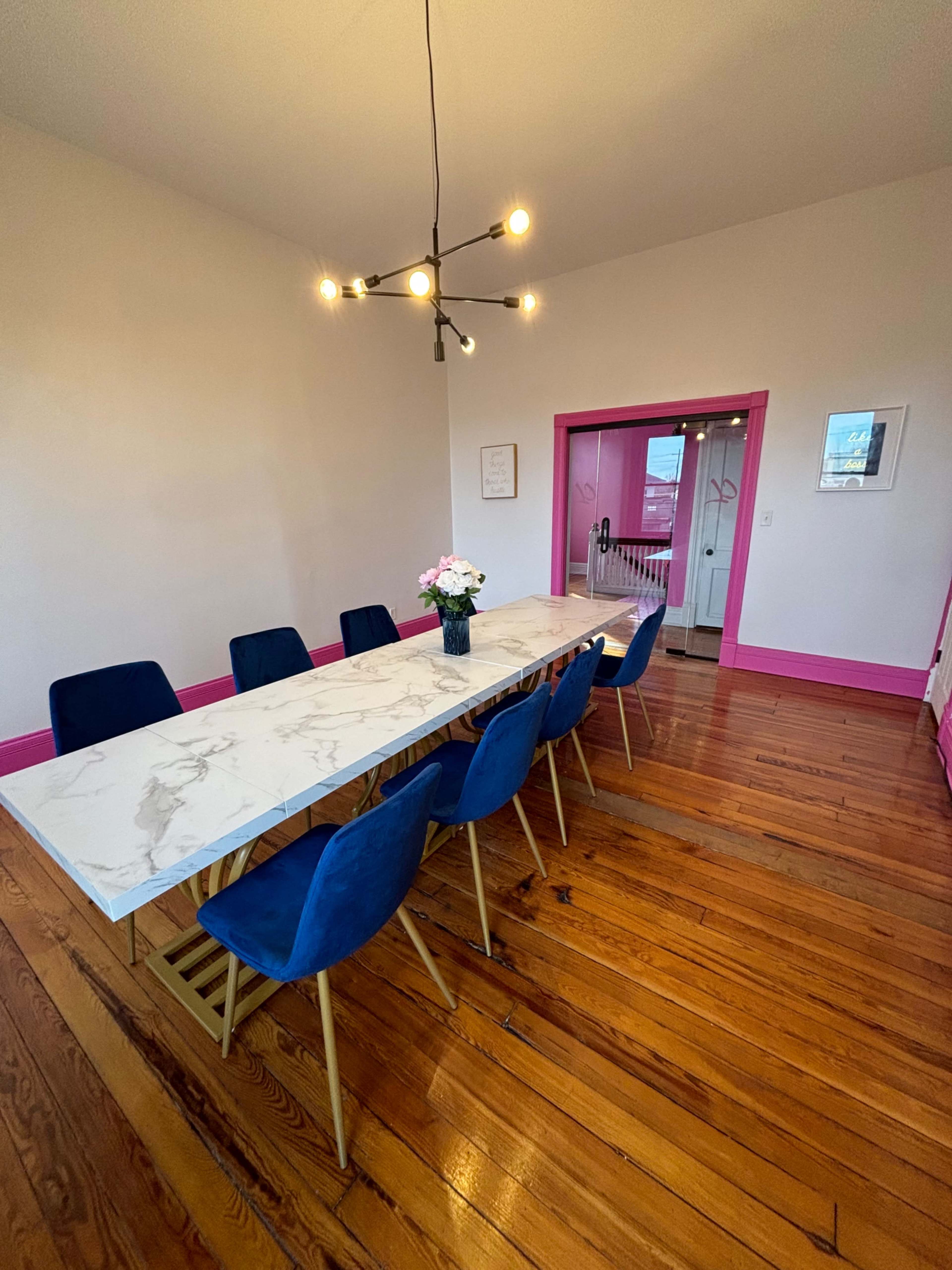 A long marble dining table with blue chairs is arranged in a room with hardwood floors and pink-accented walls.