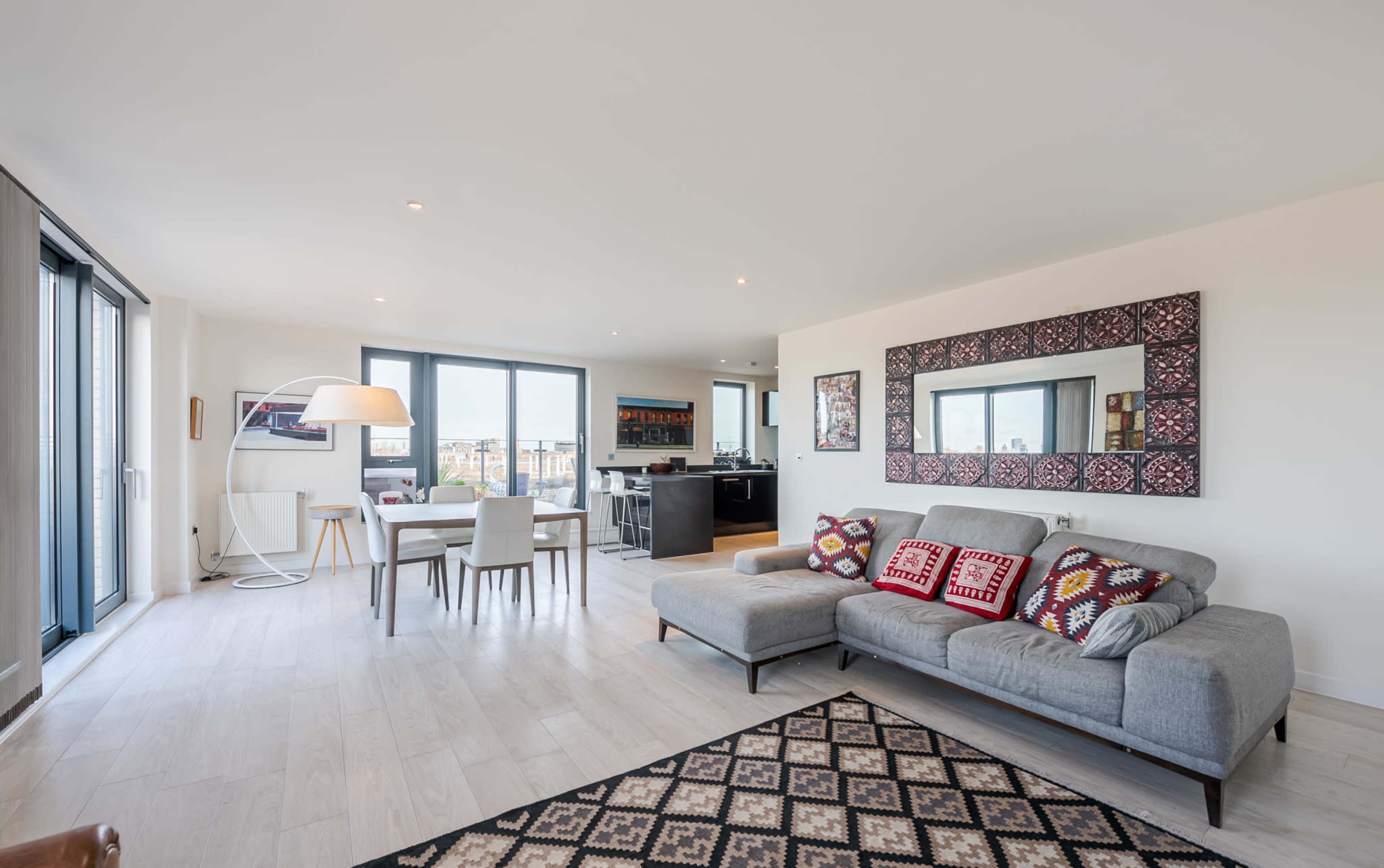 The image shows a modern living area featuring a gray sofa, a patterned rug, a dining table with chairs, and large windows that provide a view of the outside.