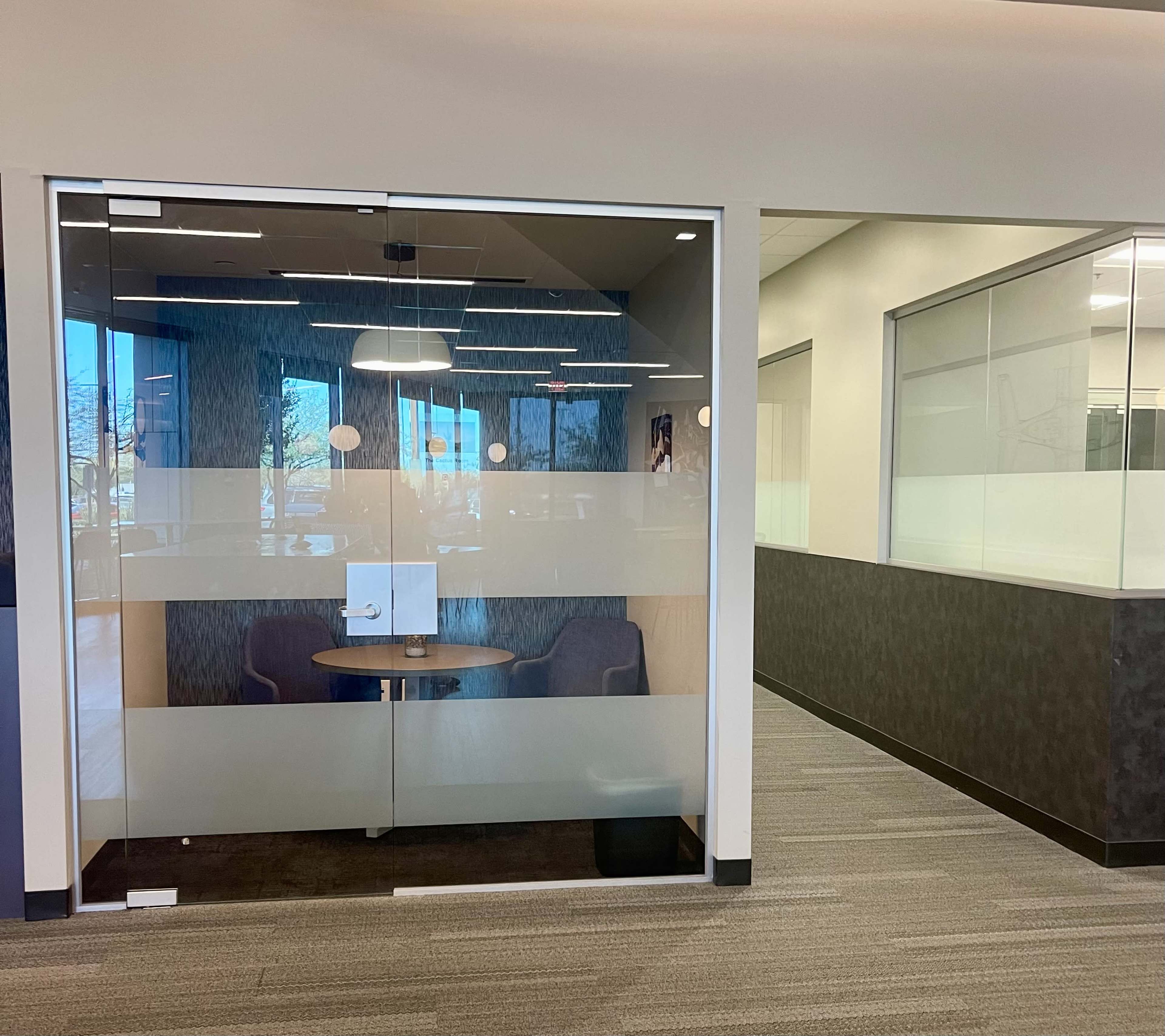 A glass-enclosed meeting room with a round table and two chairs, located in a modern office space.