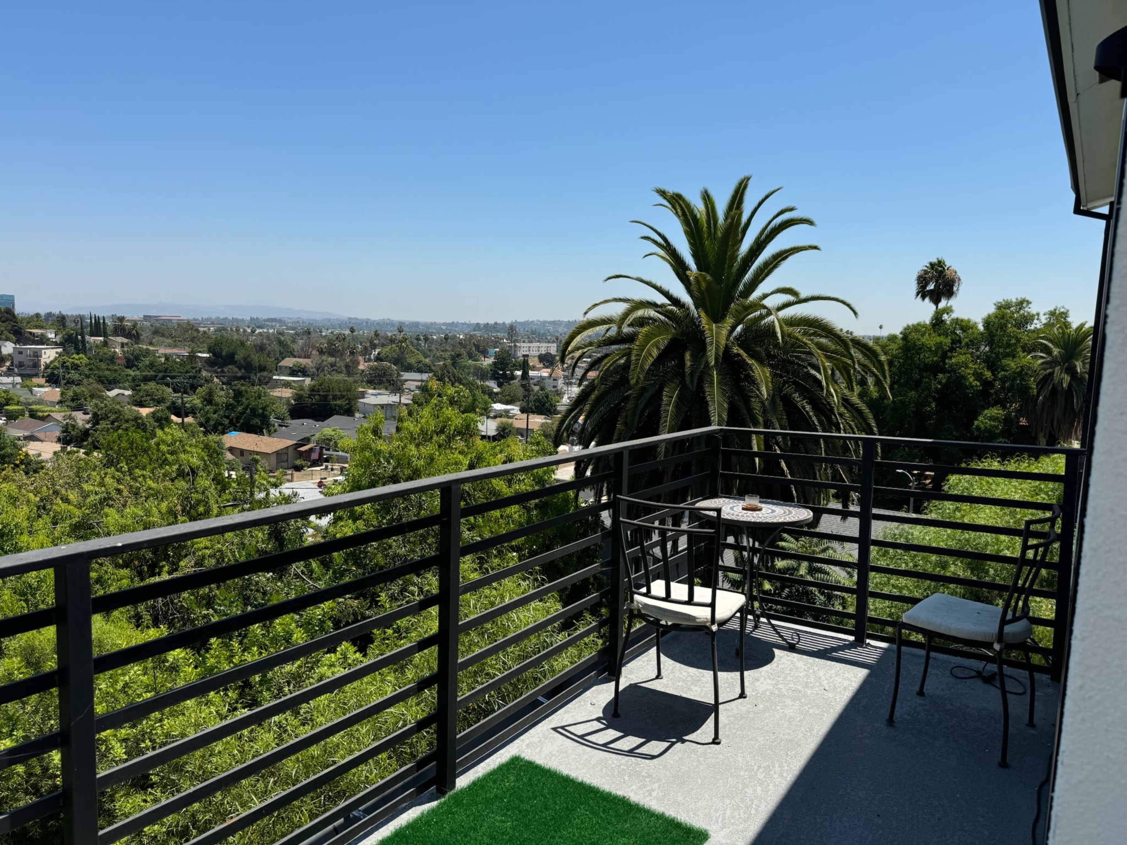A balcony with a small table and chairs overlooks a green landscape dotted with houses and palm trees under a clear blue sky.