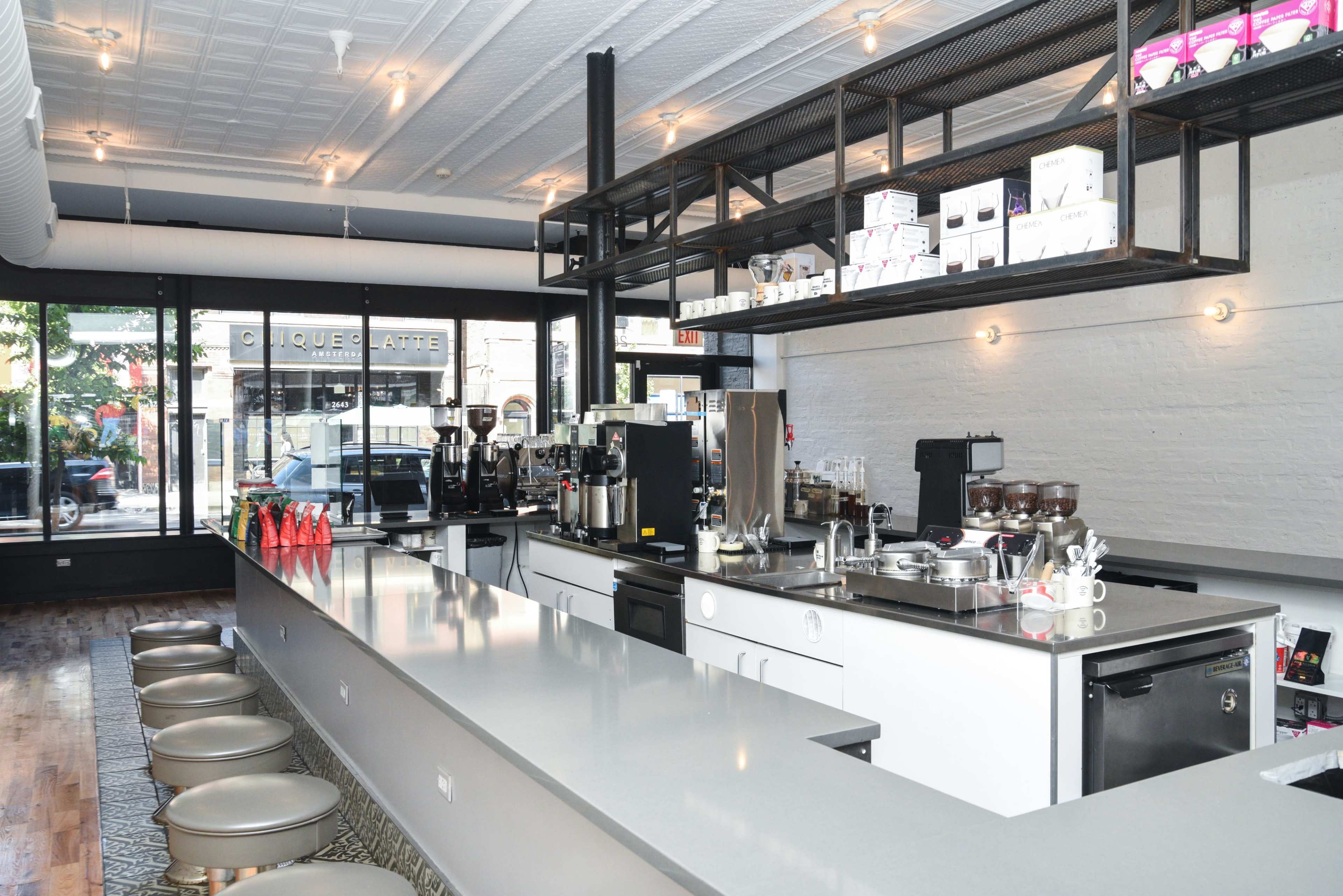 A modern, minimalist café interior featuring a long counter with coffee machines and seating in front of large windows.