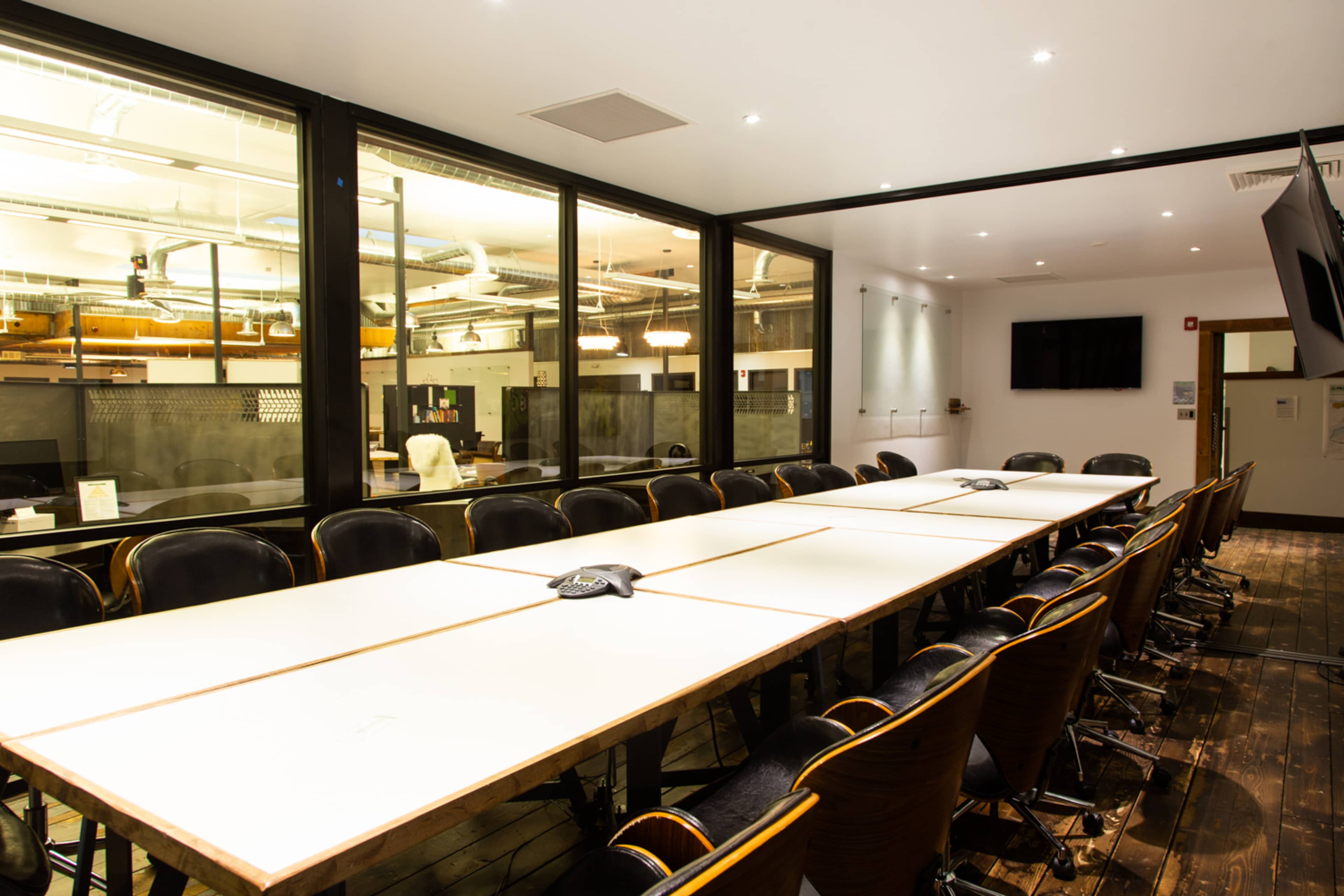 A large conference room with a long table surrounded by black office chairs, featuring glass walls and modern lighting.
