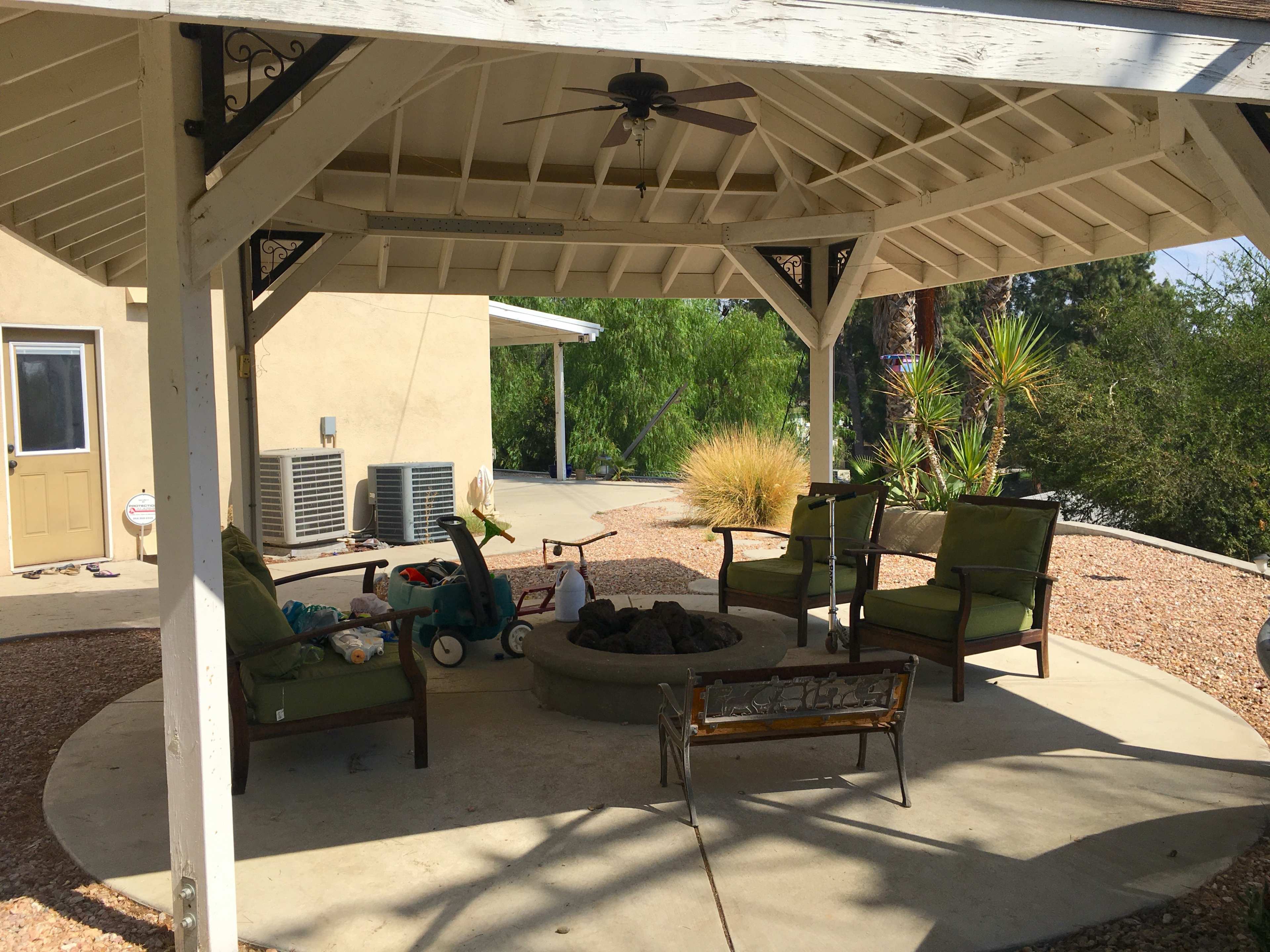 A covered patio features a circular fire pit surrounded by green chairs and a small table, with a house and vegetation visible in the background.