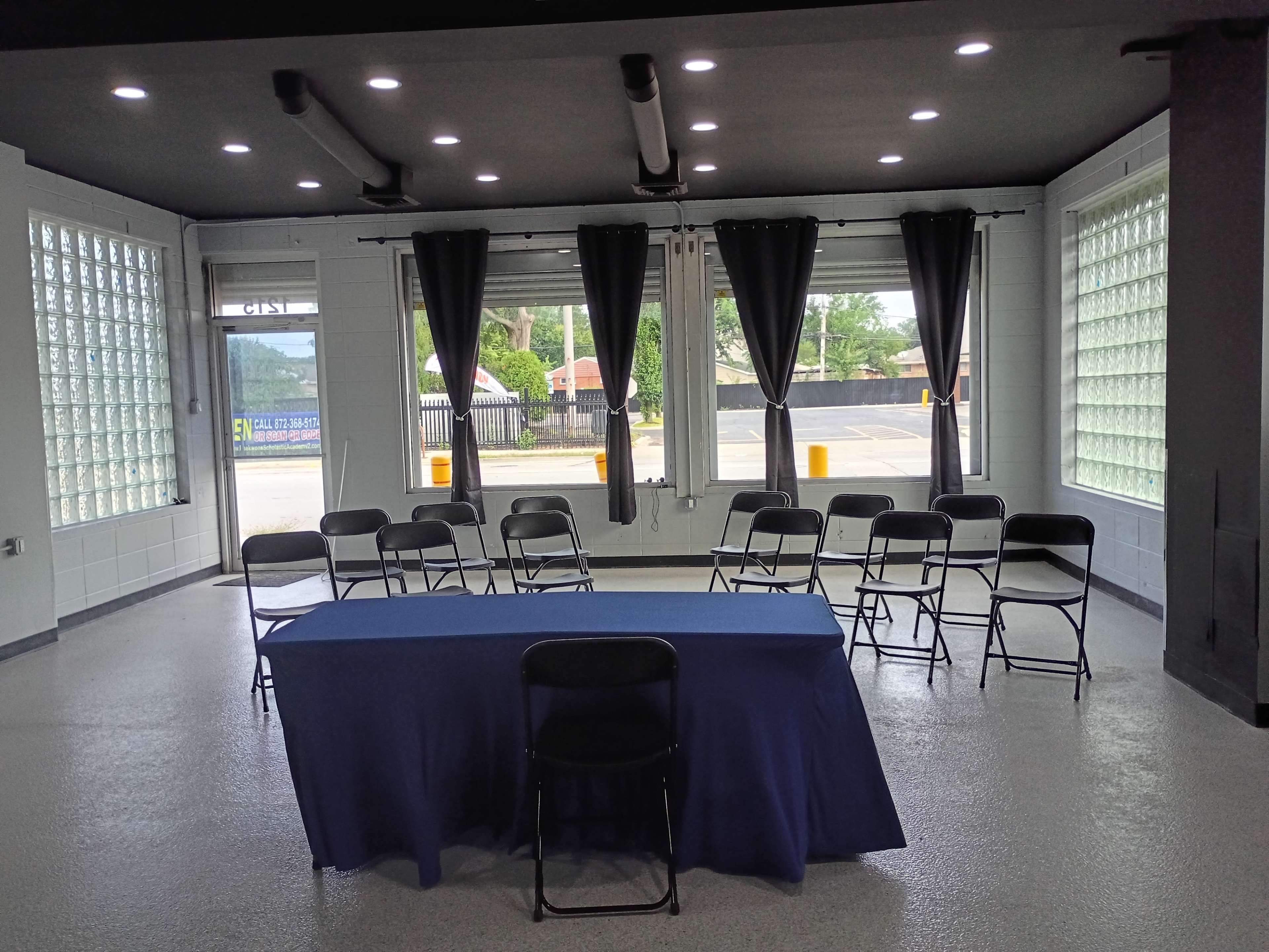 The image shows a spacious, empty room with a long blue table at the center, surrounded by black folding chairs and large windows covered by blackout curtains.