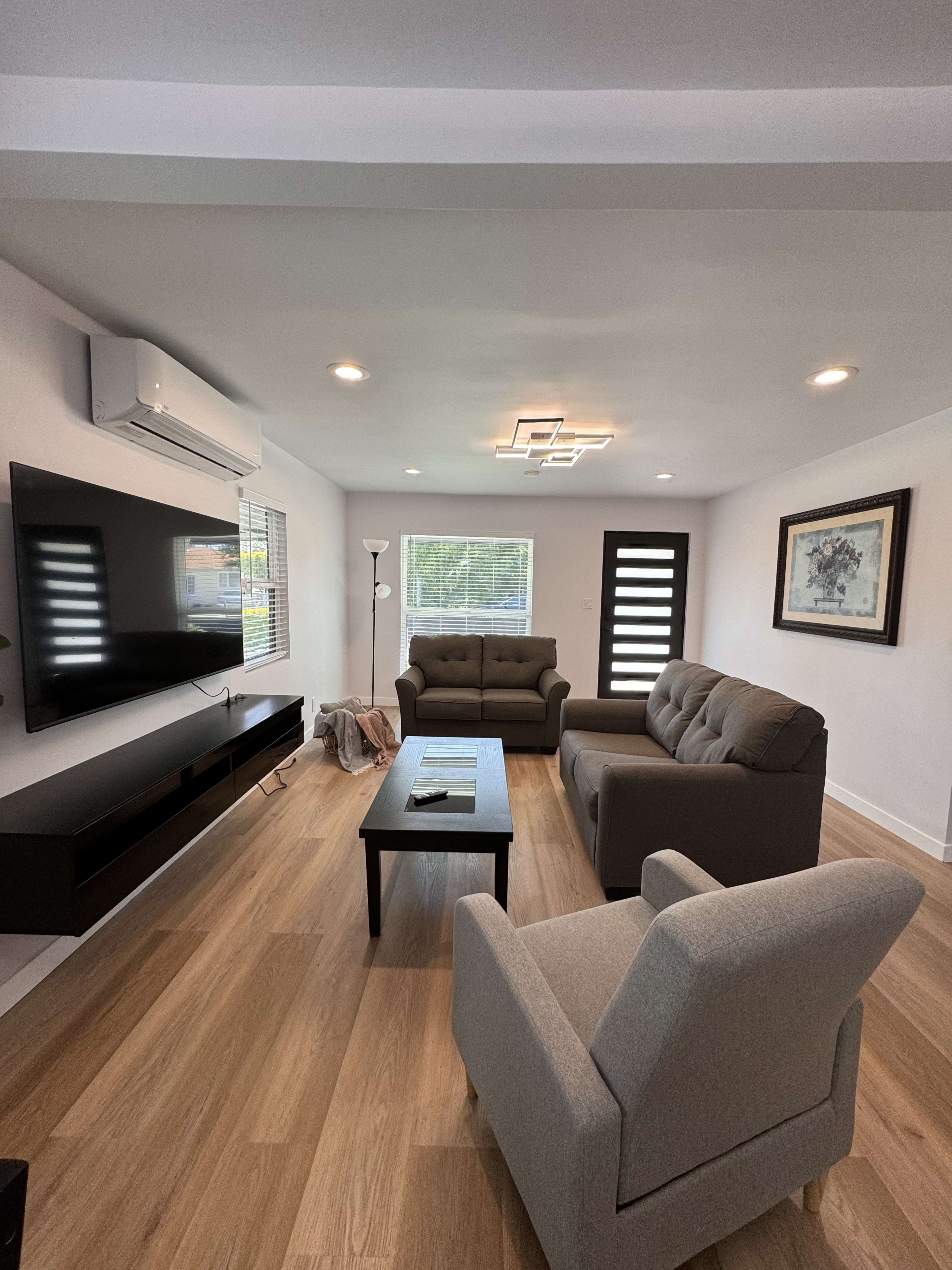 A modern living room with two sofas, a coffee table, a TV mounted on the wall, and large windows allowing natural light.