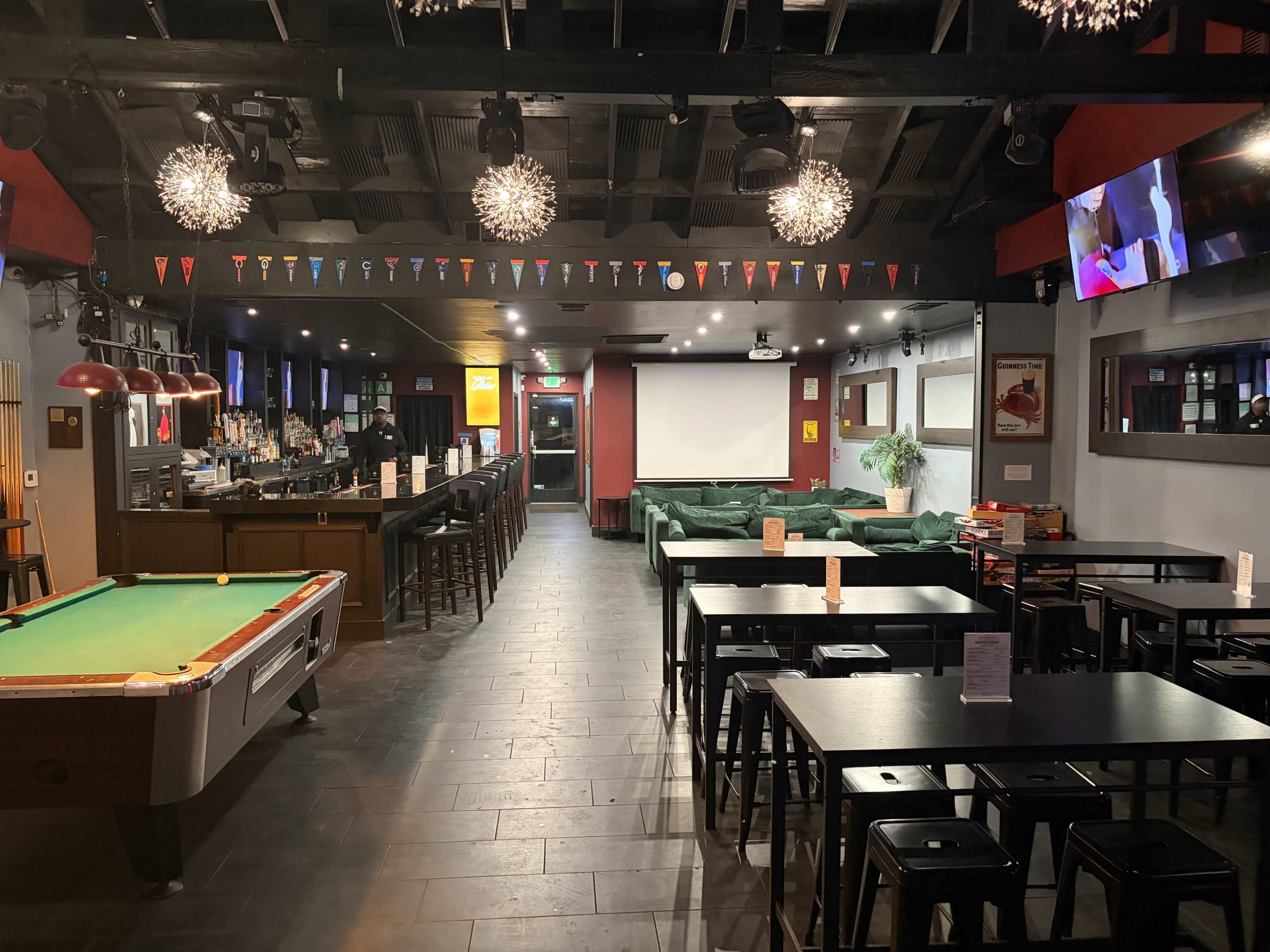 The image shows the interior of a bar or pub with a pool table, tables, and a bar area visible, along with a large screen at one end and decorative lighting.