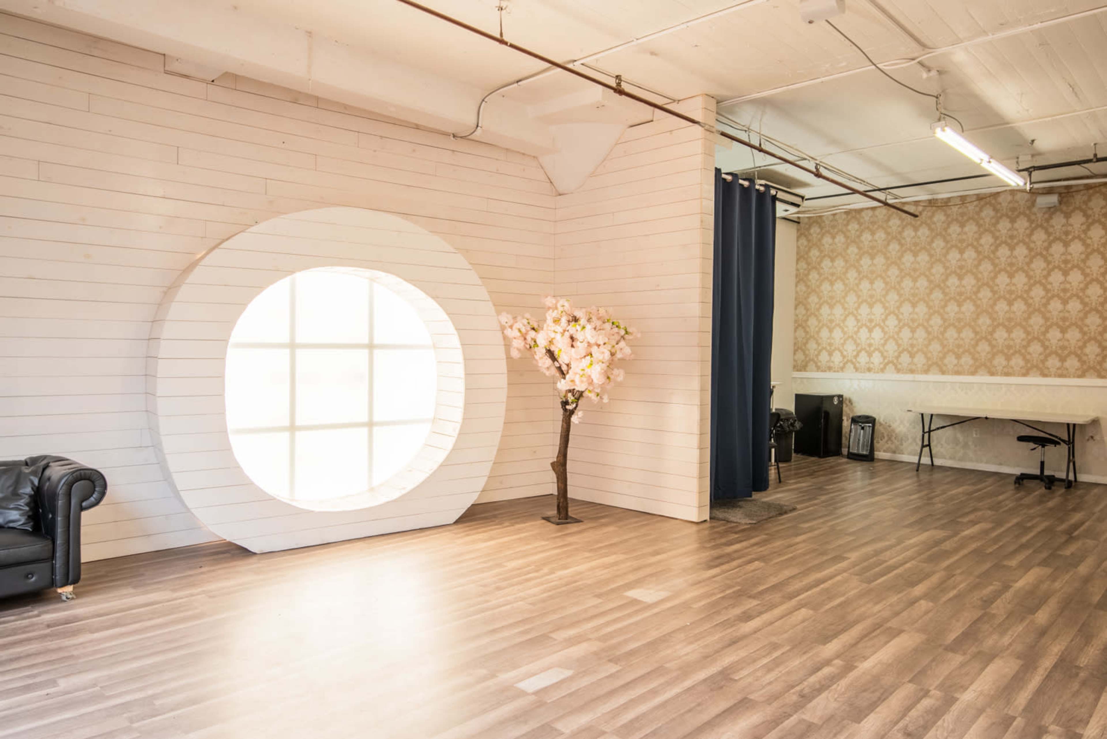A spacious, brightly lit studio featuring a large circular window, a decorative cherry blossom tree, and an area with a table and chairs.