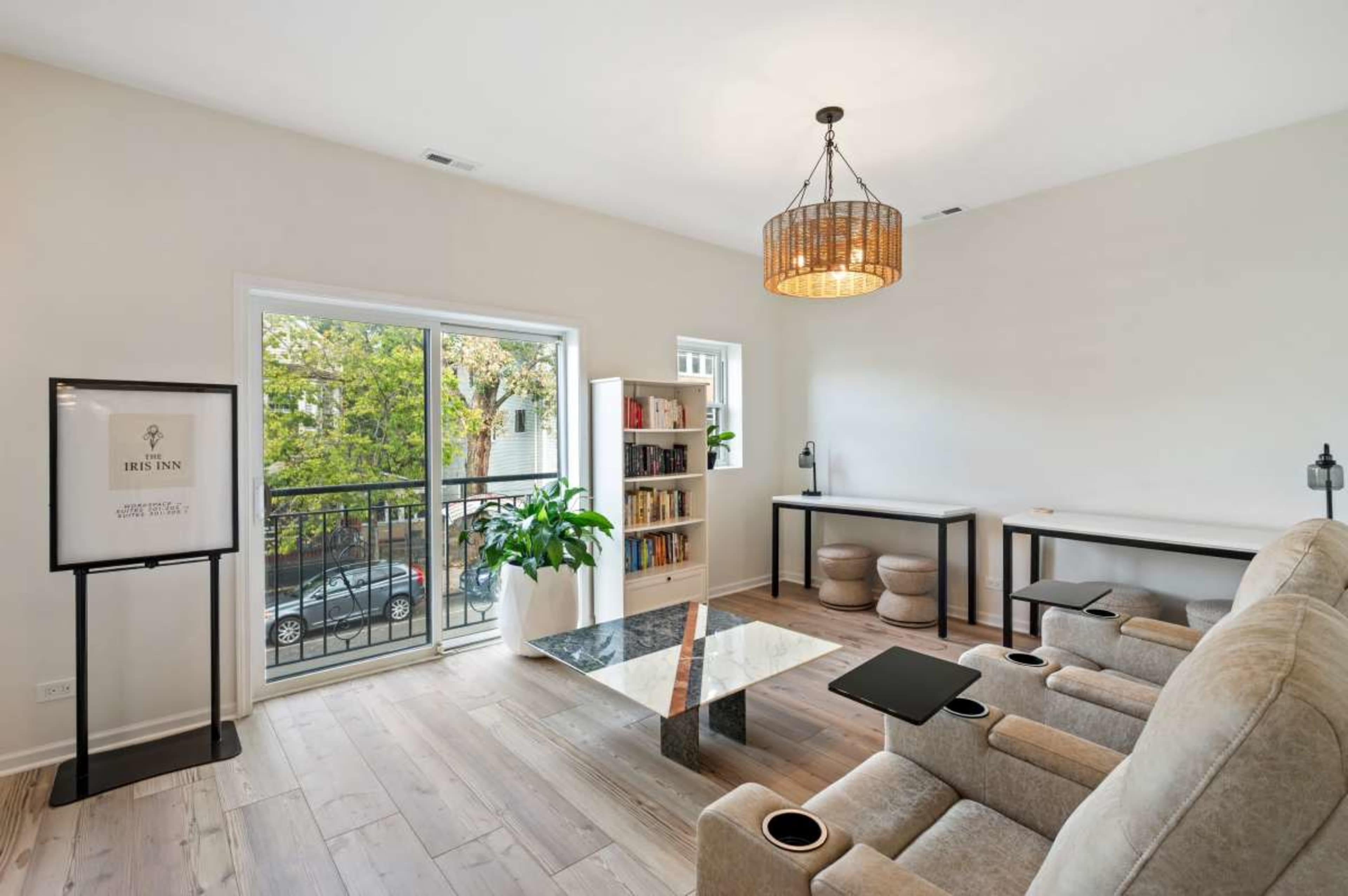 The room features a light-colored design with a seating area, a coffee table, a bookshelf, and large windows overlooking a balcony.