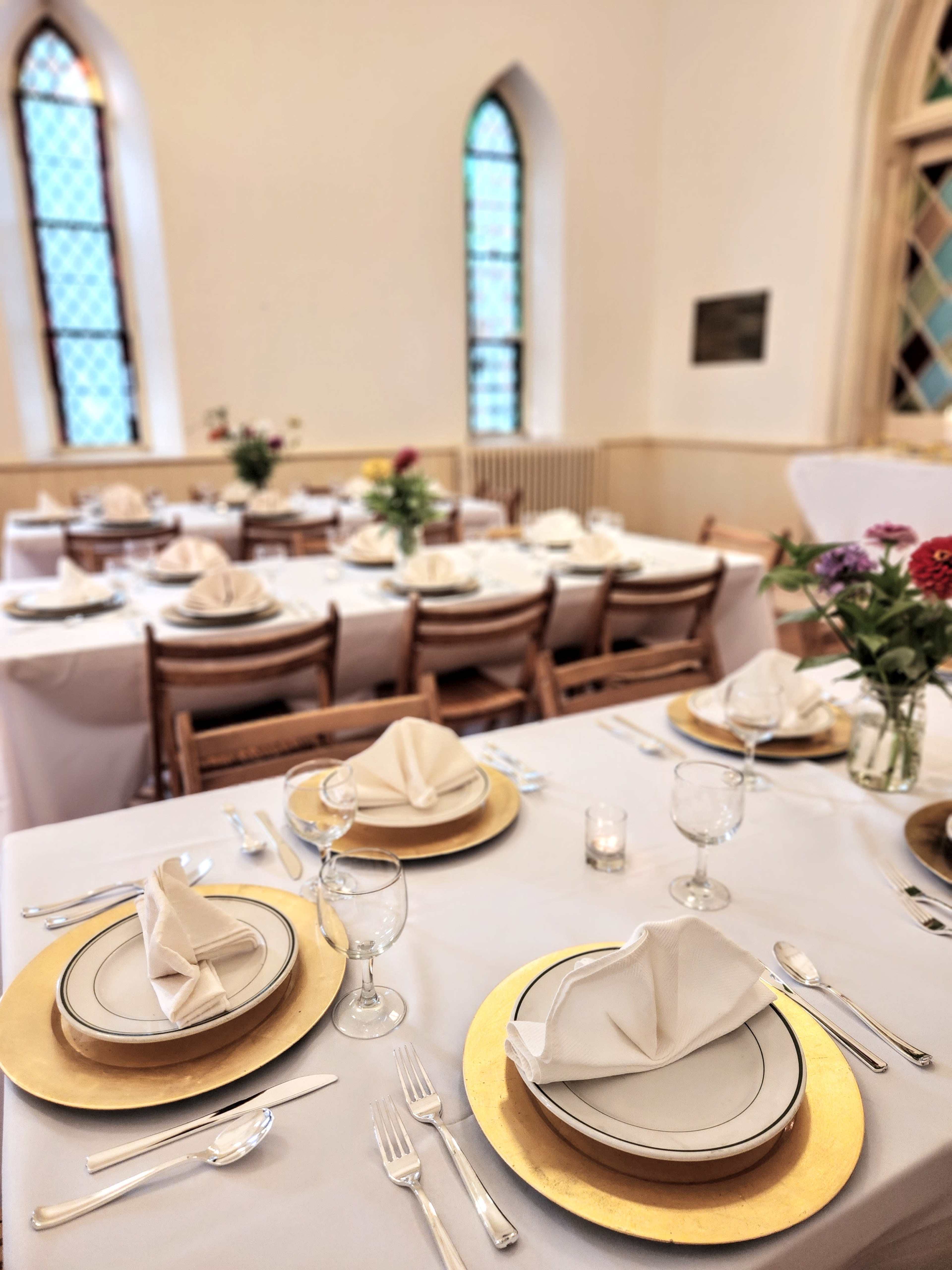 A dining area is set up with elegantly arranged tables, each featuring plates, glasses, and folded napkins, with flowers in vases as centerpieces.