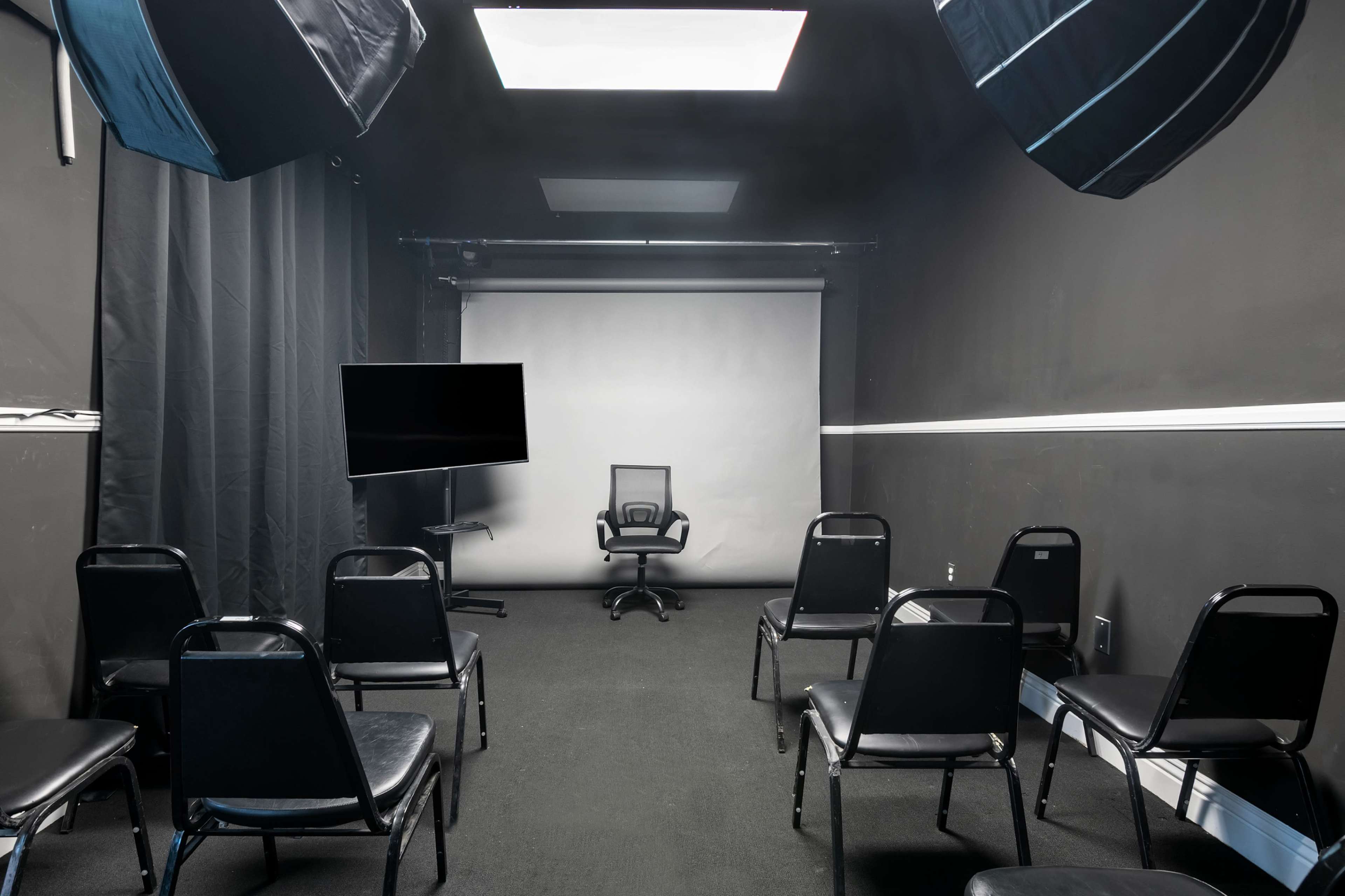 A small, dimly lit room features a center chair facing a blank screen, surrounded by several black chairs and equipped with lighting setups.