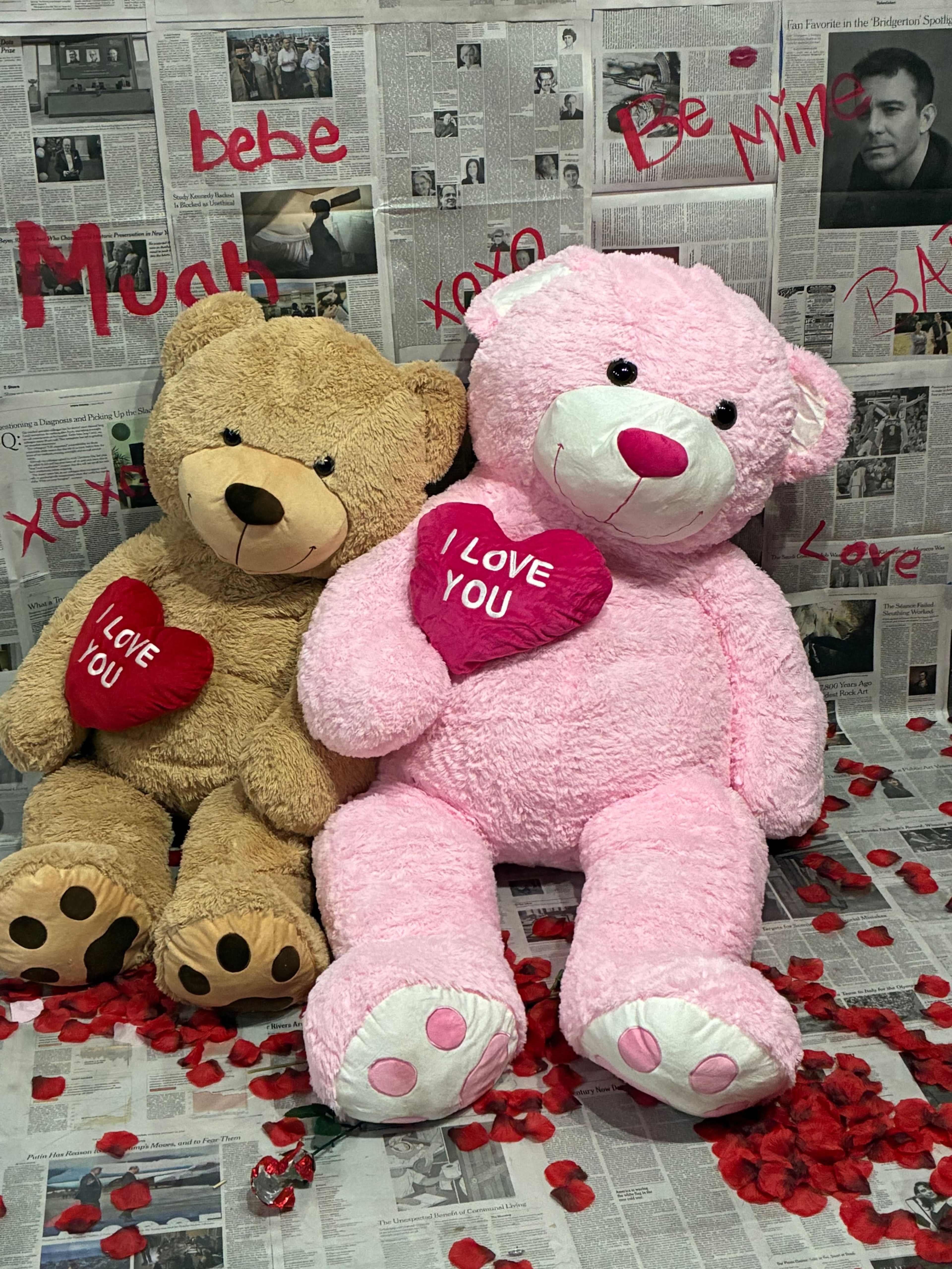 The image features two large stuffed bears, one brown and one pink, sitting among rose petals on a background of newspaper clippings with various romantic phrases.