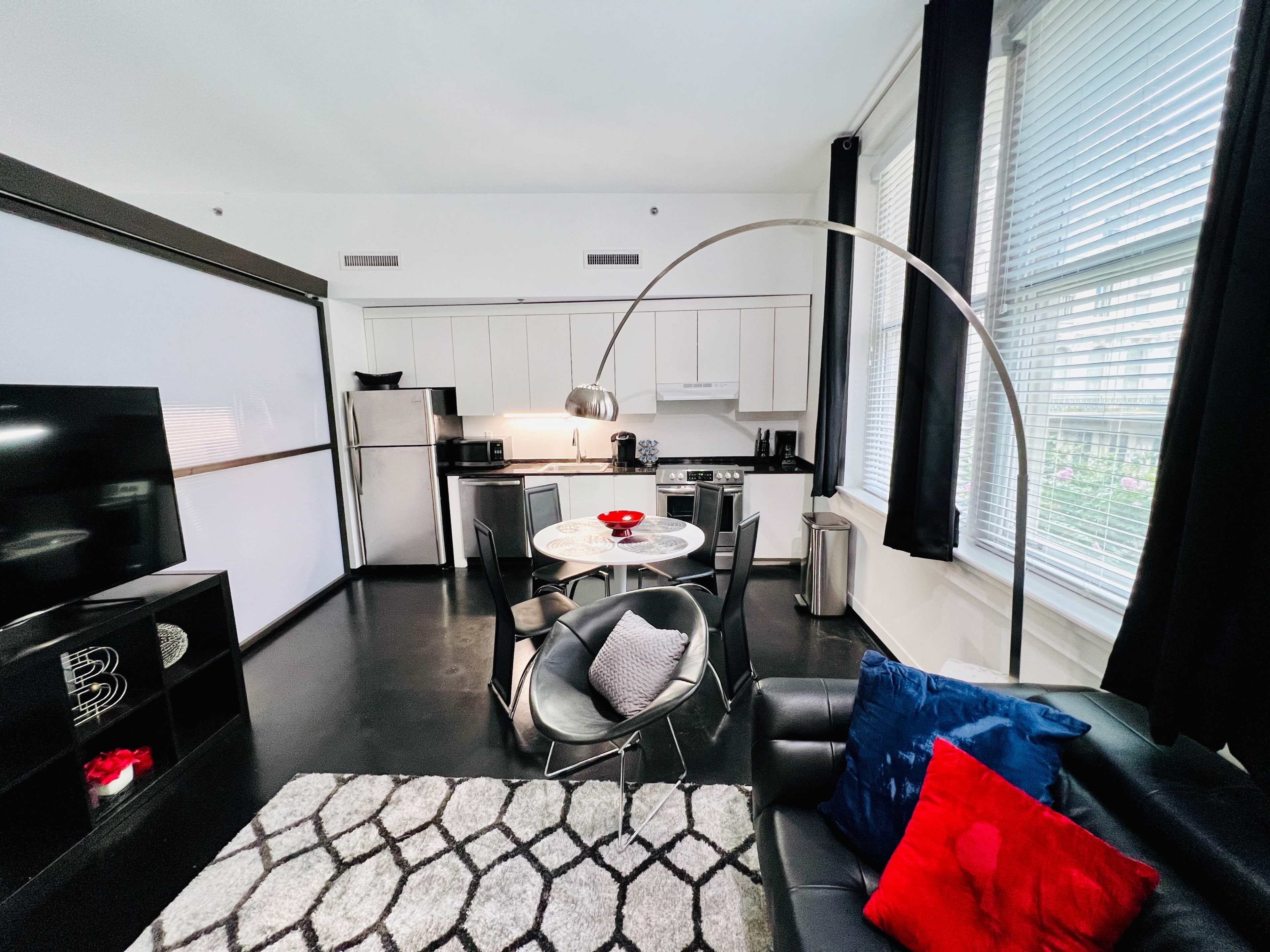 A modern kitchen and dining area feature a circular table with chairs, a sleek couch, and large windows allowing natural light.